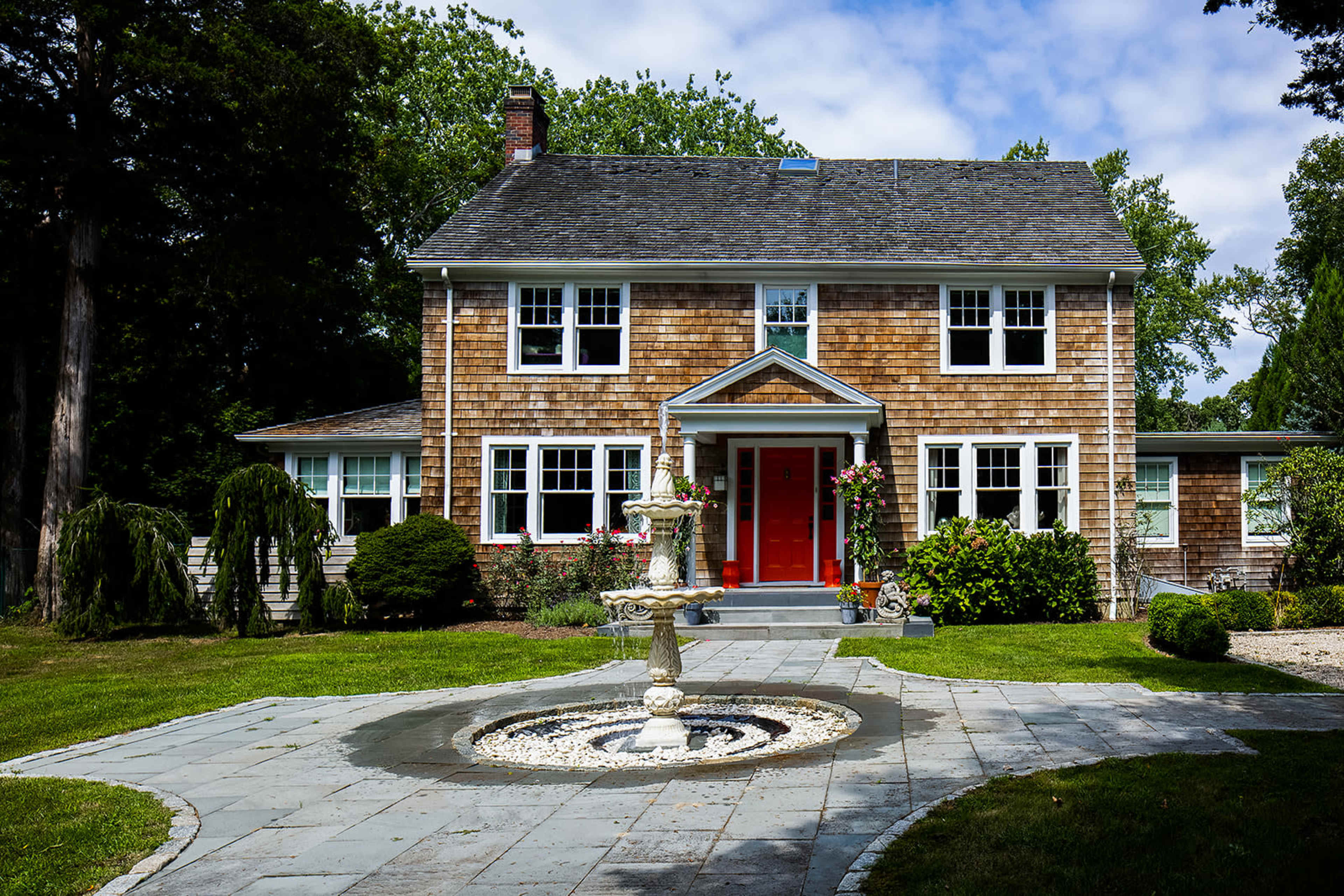 A two-story wooden house with a red front door, surrounded by a landscaped garden and a stone fountain in the foreground.