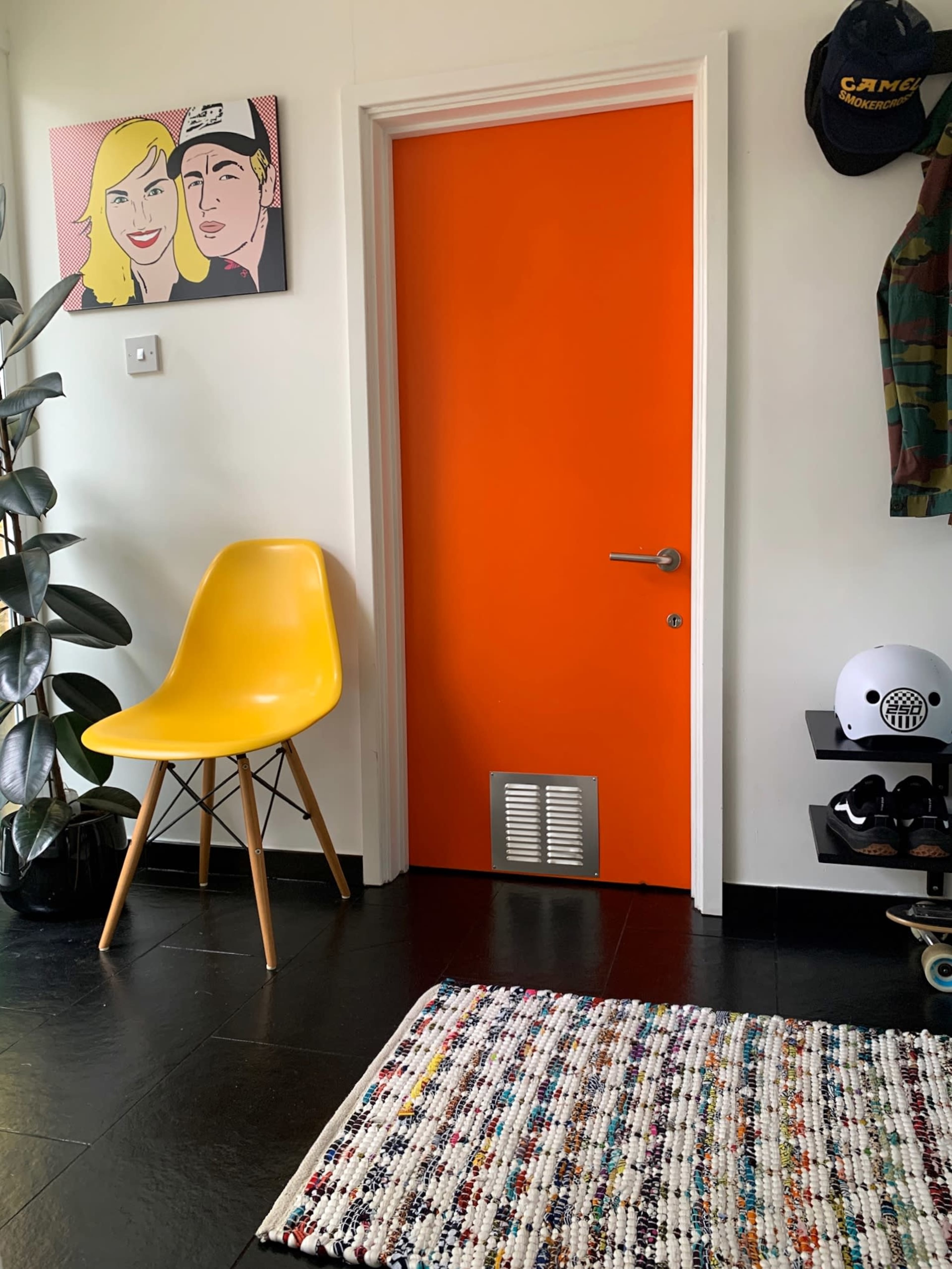 An orange door in a room with a yellow chair, a colorful textured rug, and a wall featuring a framed artwork of two people.