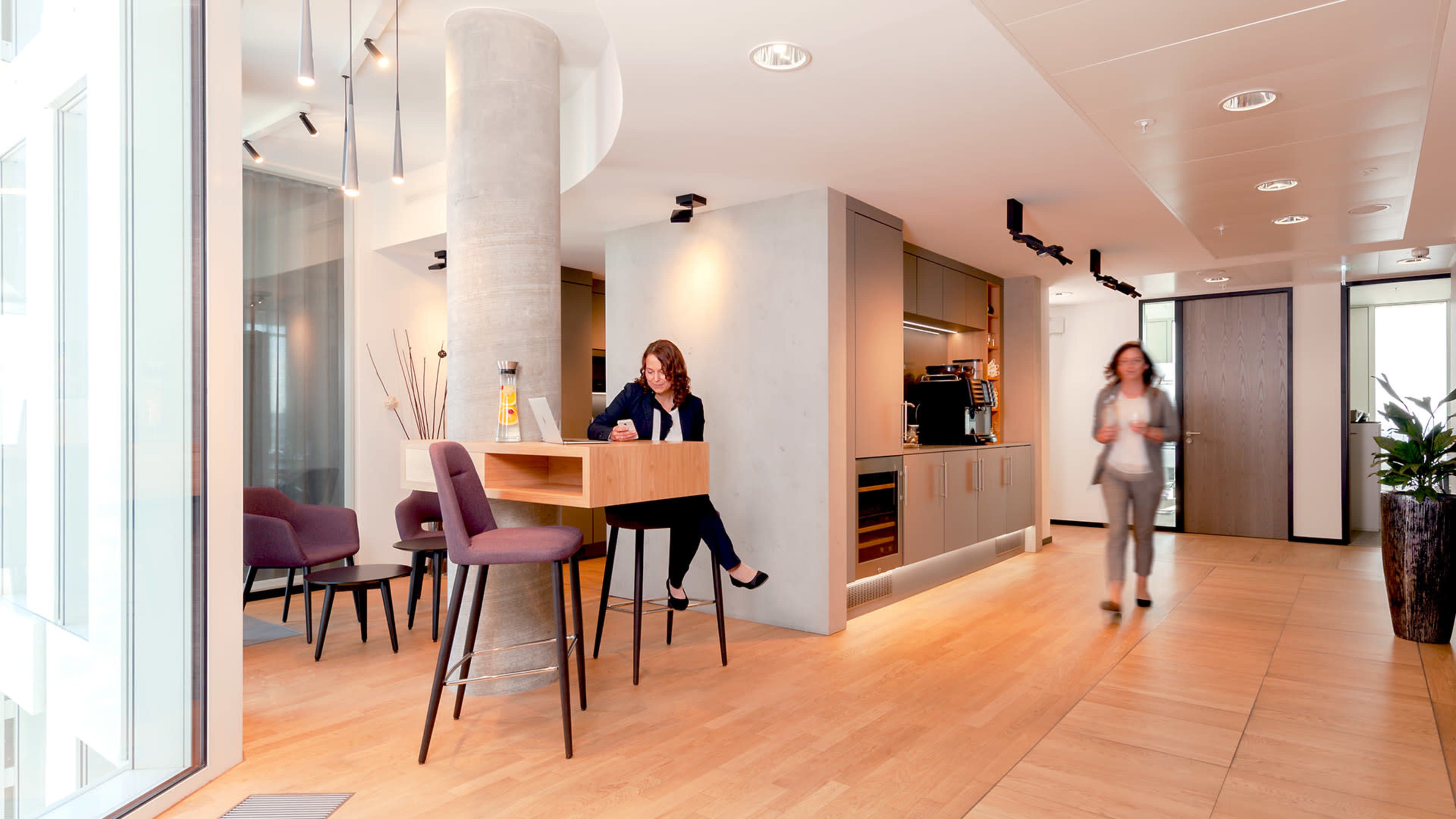 The image shows a modern office interior with a kitchenette and seating area, featuring a woman sitting at a high table and another woman walking through the space.