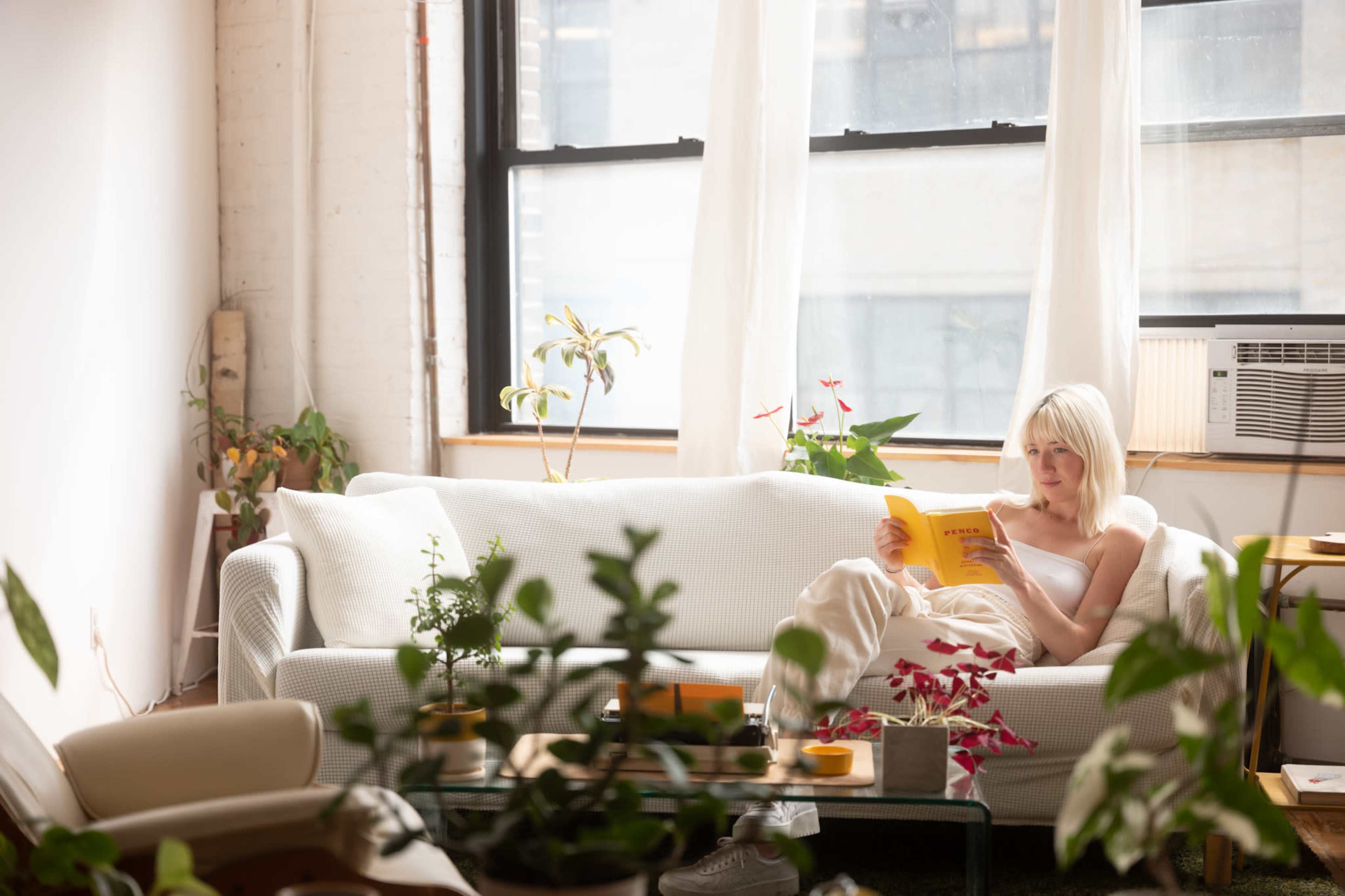 A woman sits on a white couch reading a book in a sunlit room filled with plants.