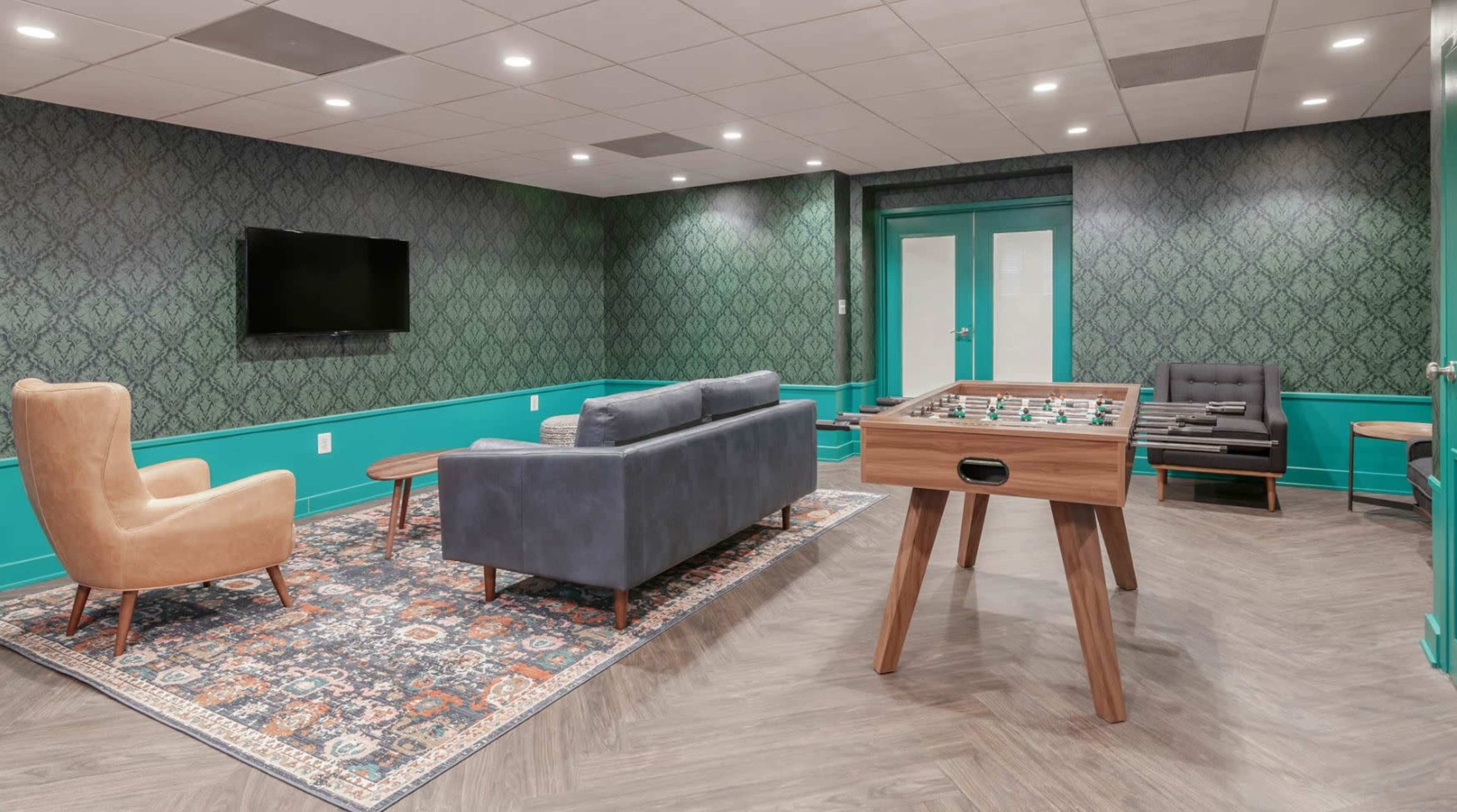 A stylish lounge area featuring a foosball table, a gray sofa, a brown armchair, and patterned wallpaper.