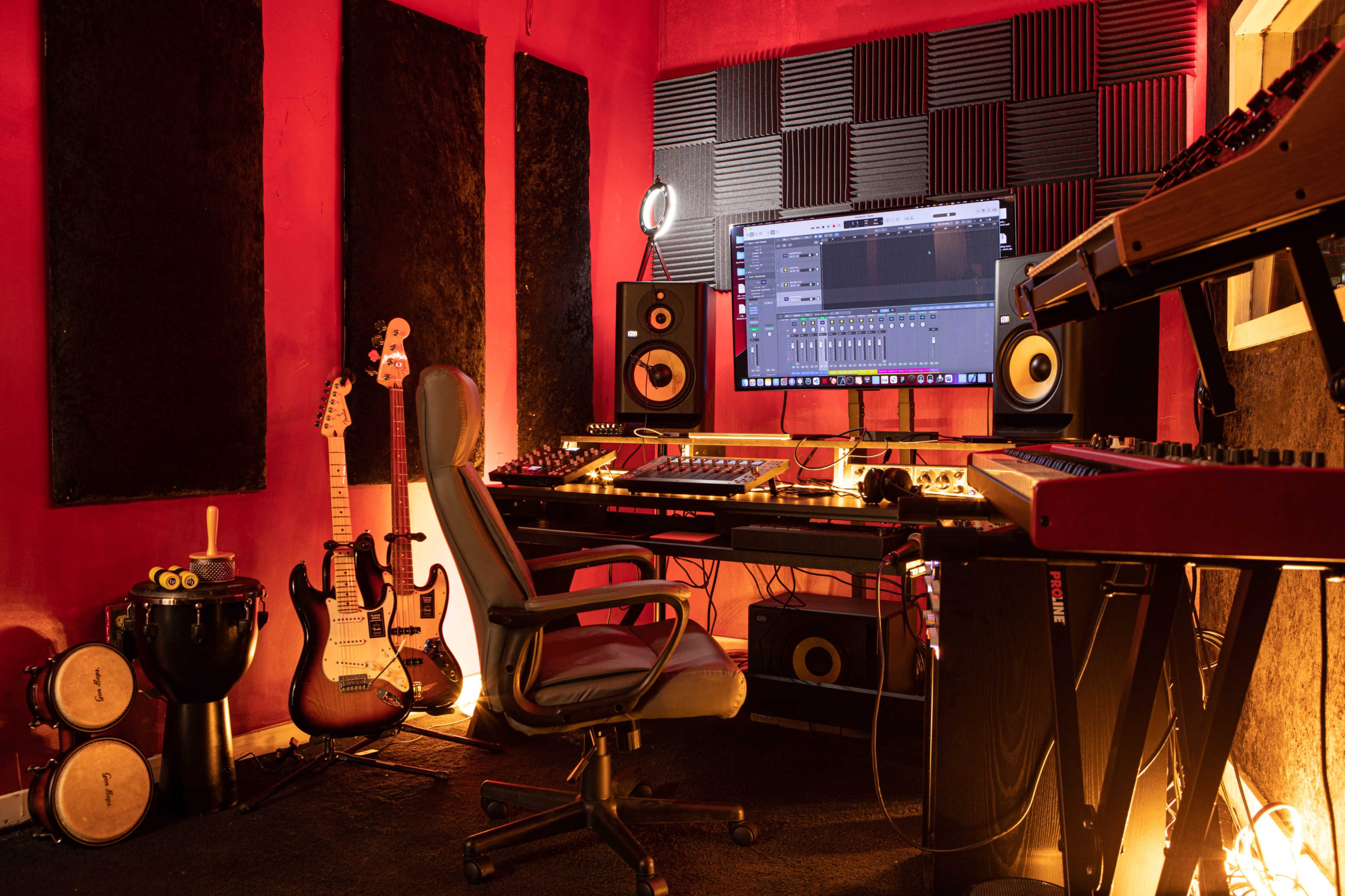 A music production studio with a large monitor, various musical instruments, a chair, and soundproof panels on the walls.