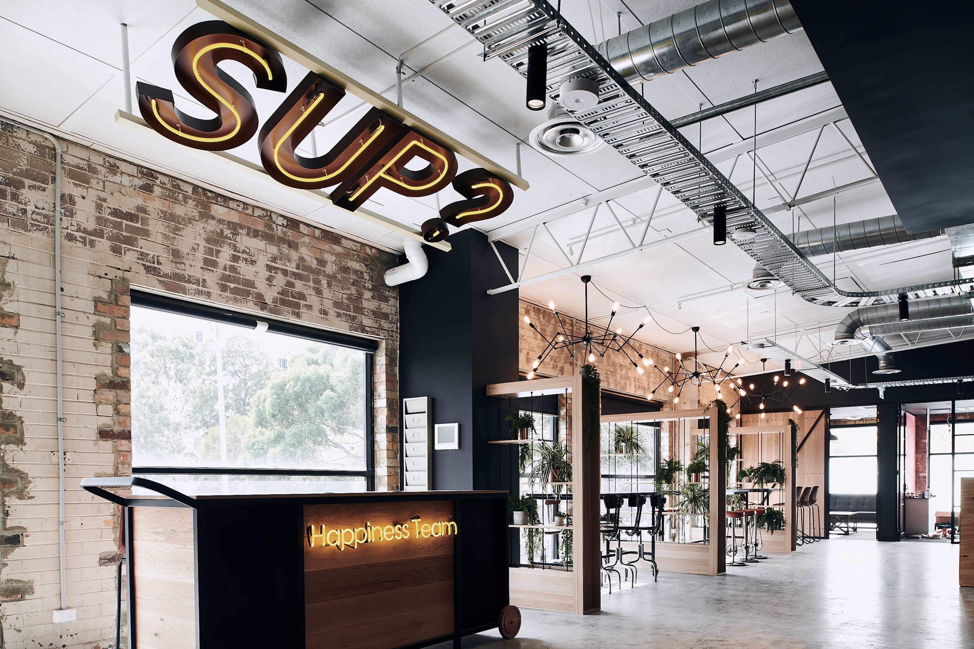 The image shows a modern office space with a reception area featuring a wooden desk labeled "Happiness Team" and a large illuminated sign that reads "SUP?" on the wall.