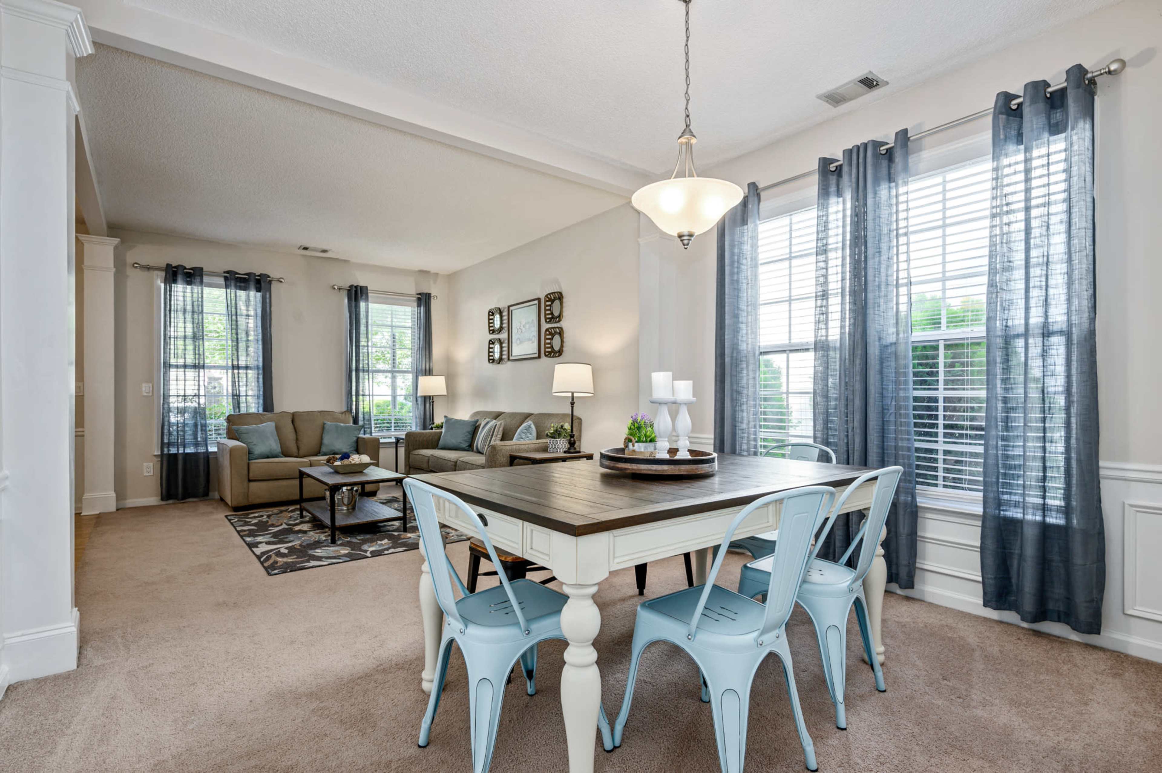 A dining area with a light wooden table and blue metal chairs is situated next to a living space featuring a sofa and coffee table, illuminated by natural light from large windows.