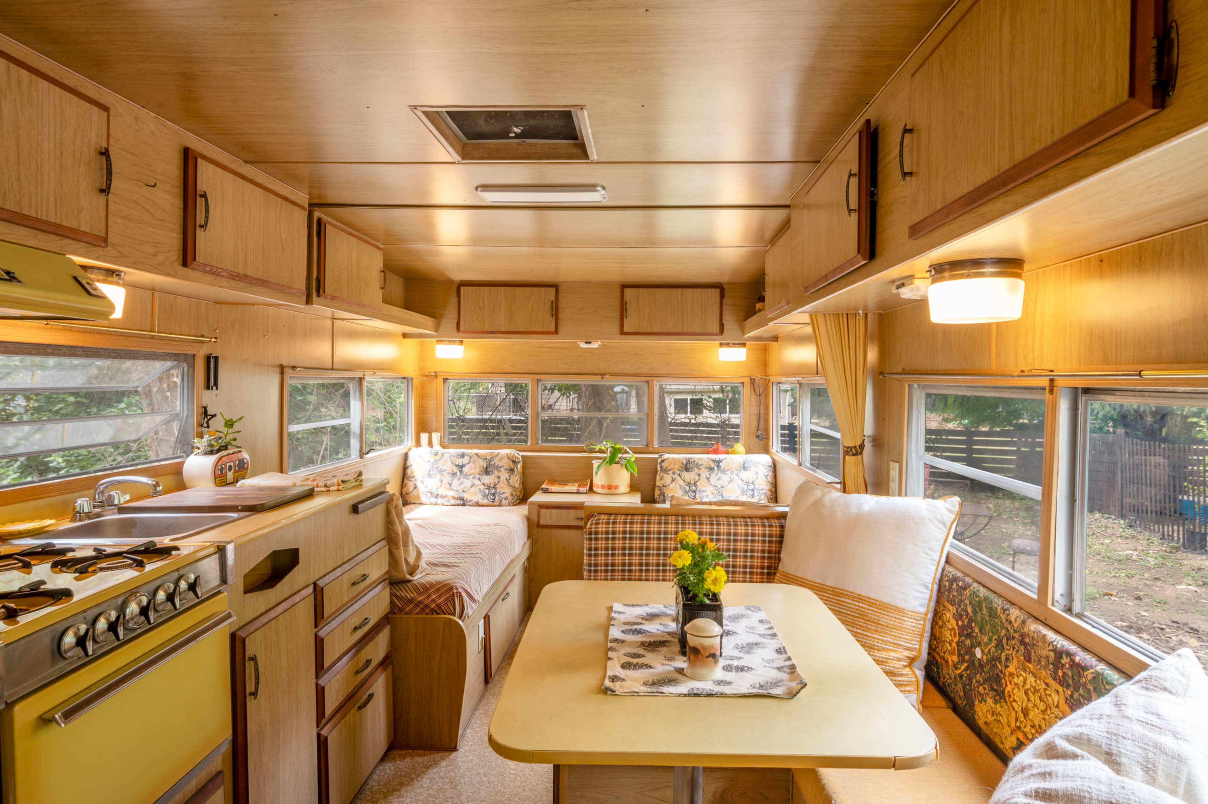 The interior of a vintage camper showcases wood-paneled walls, a kitchen area with a stove, and a dining space surrounded by seating and windows.