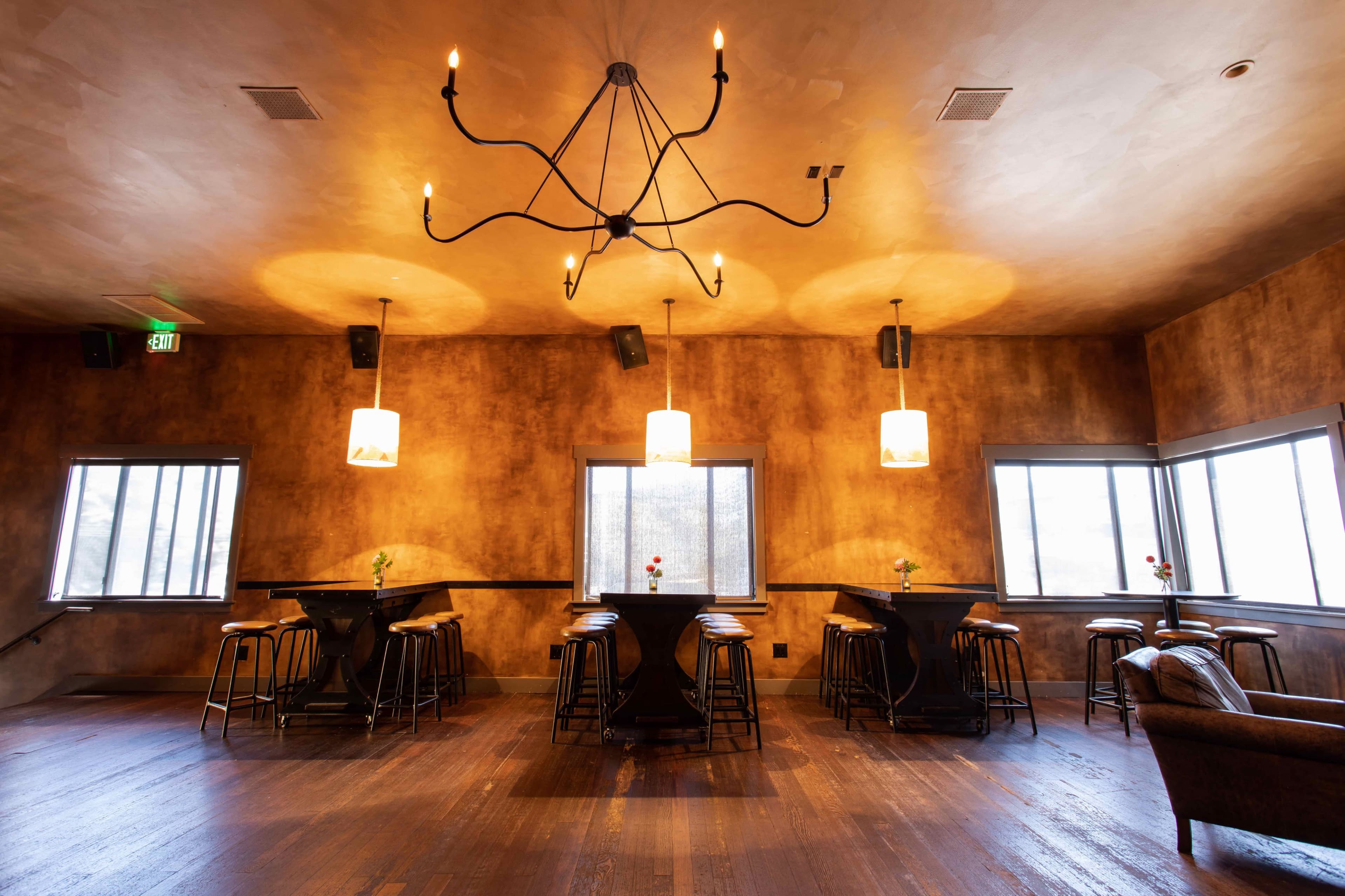 A dimly lit restaurant interior featuring a decorative chandelier, several table setups with stools, and windows allowing for natural light.
