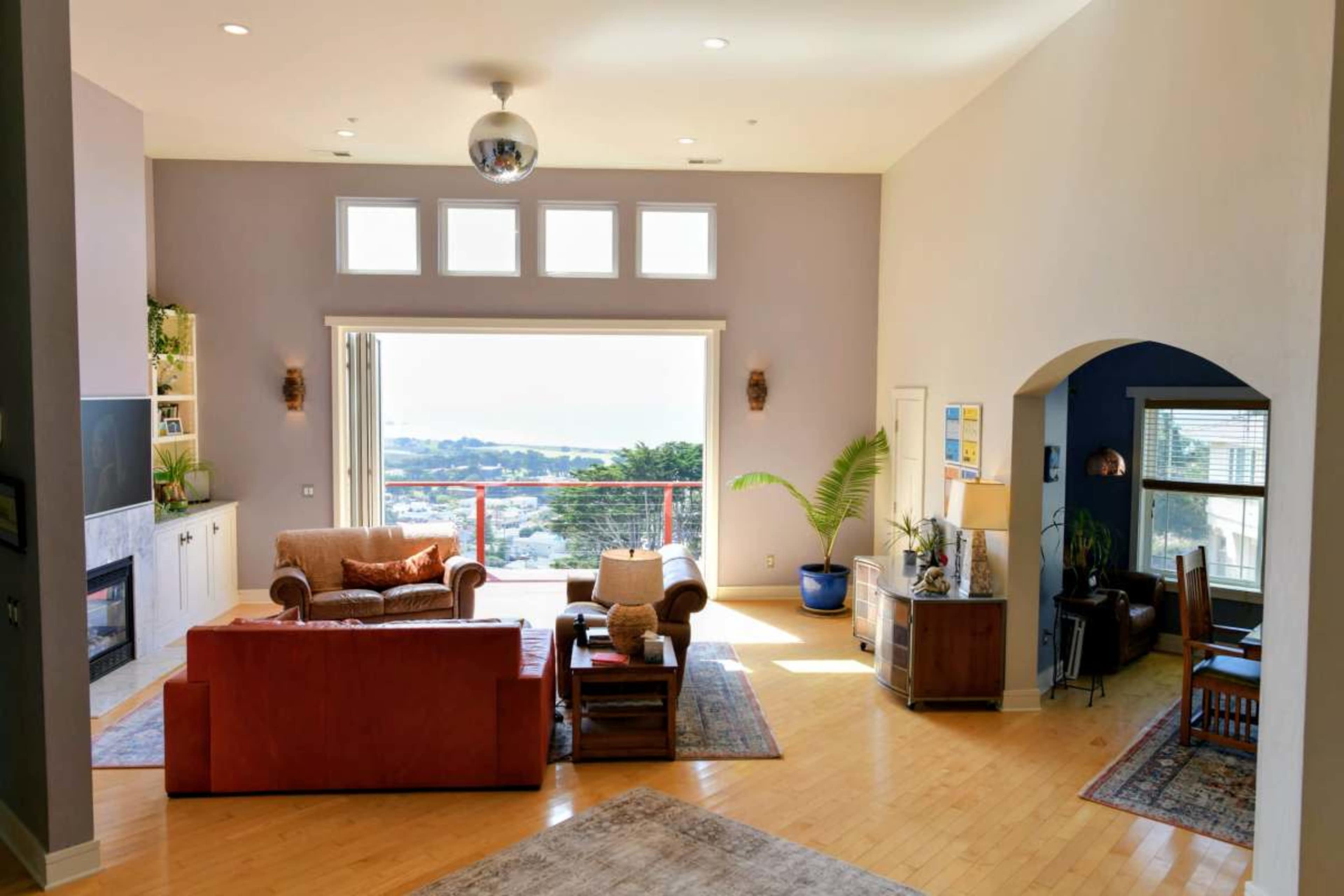 The image shows a spacious living room with a large window offering a view of the outdoors and furnishings arranged around a central seating area.