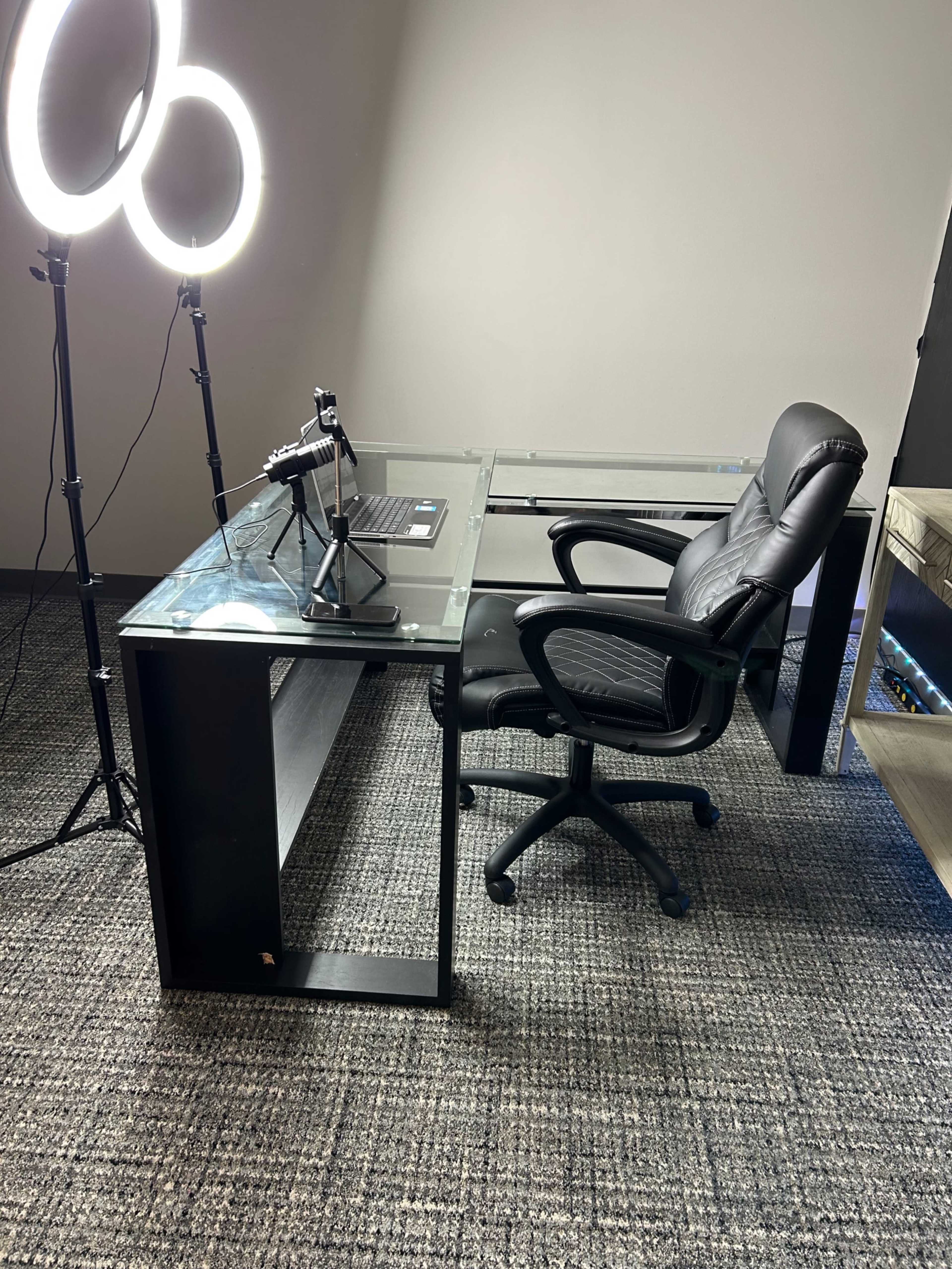 A modern glass desk with a black office chair and a ring light setup, accompanied by a microphone and a laptop.
