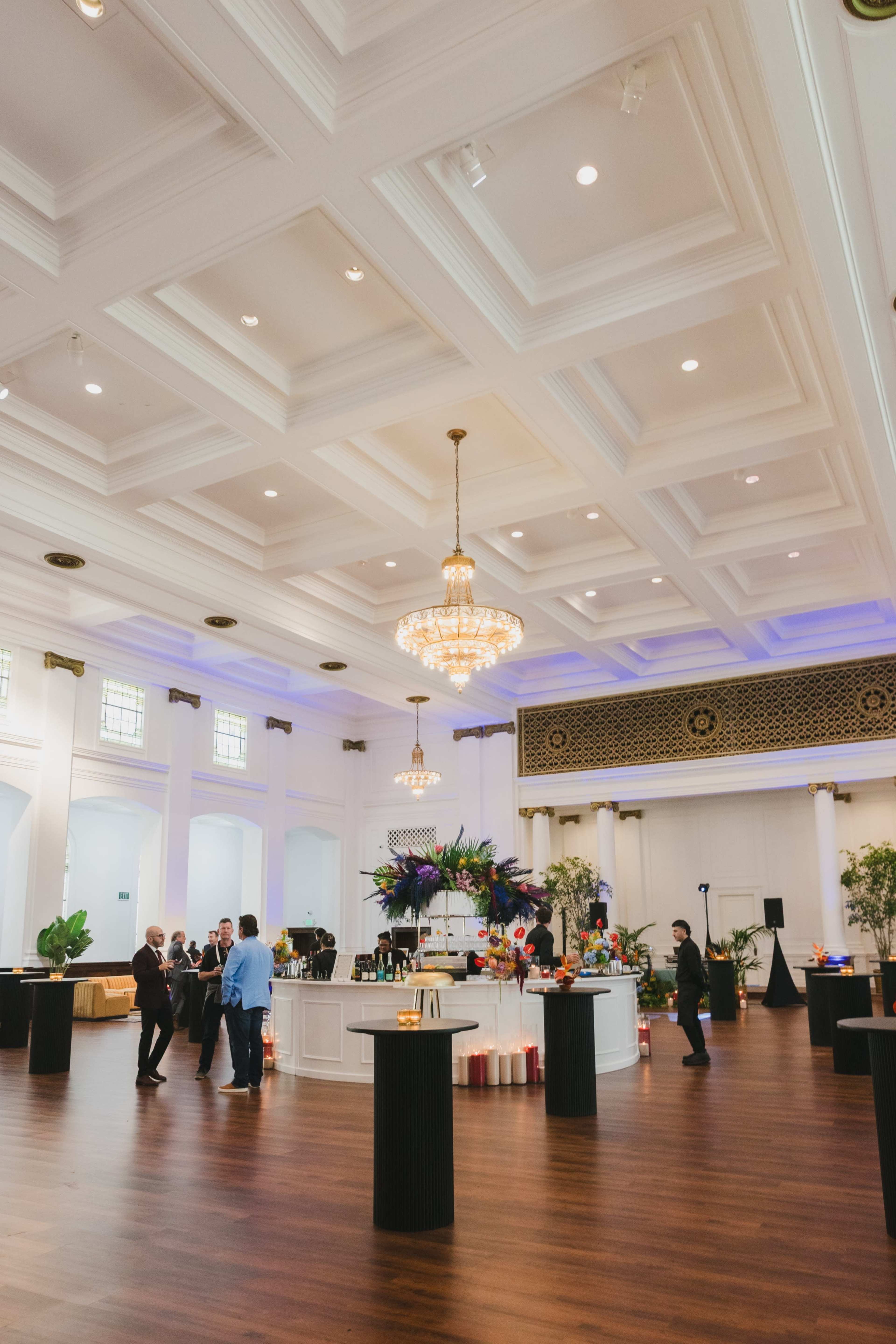 The image shows a spacious event hall with high ceilings, elegant chandeliers, and a central bar area surrounded by guests mingling.