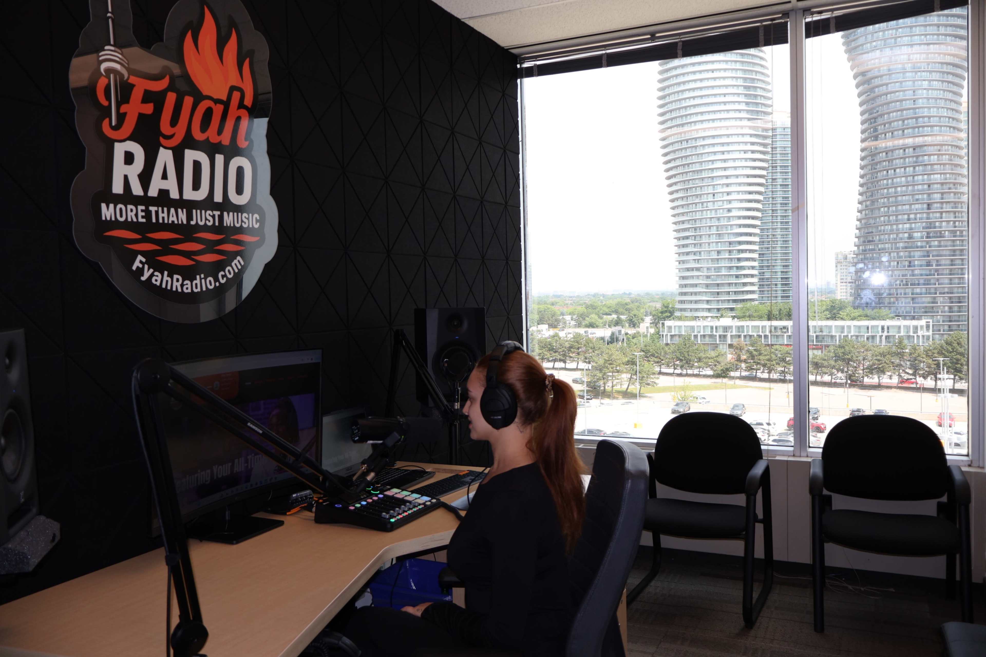 A woman sits at a radio station desk wearing headphones, with a large window showcasing modern skyscrapers in the background.