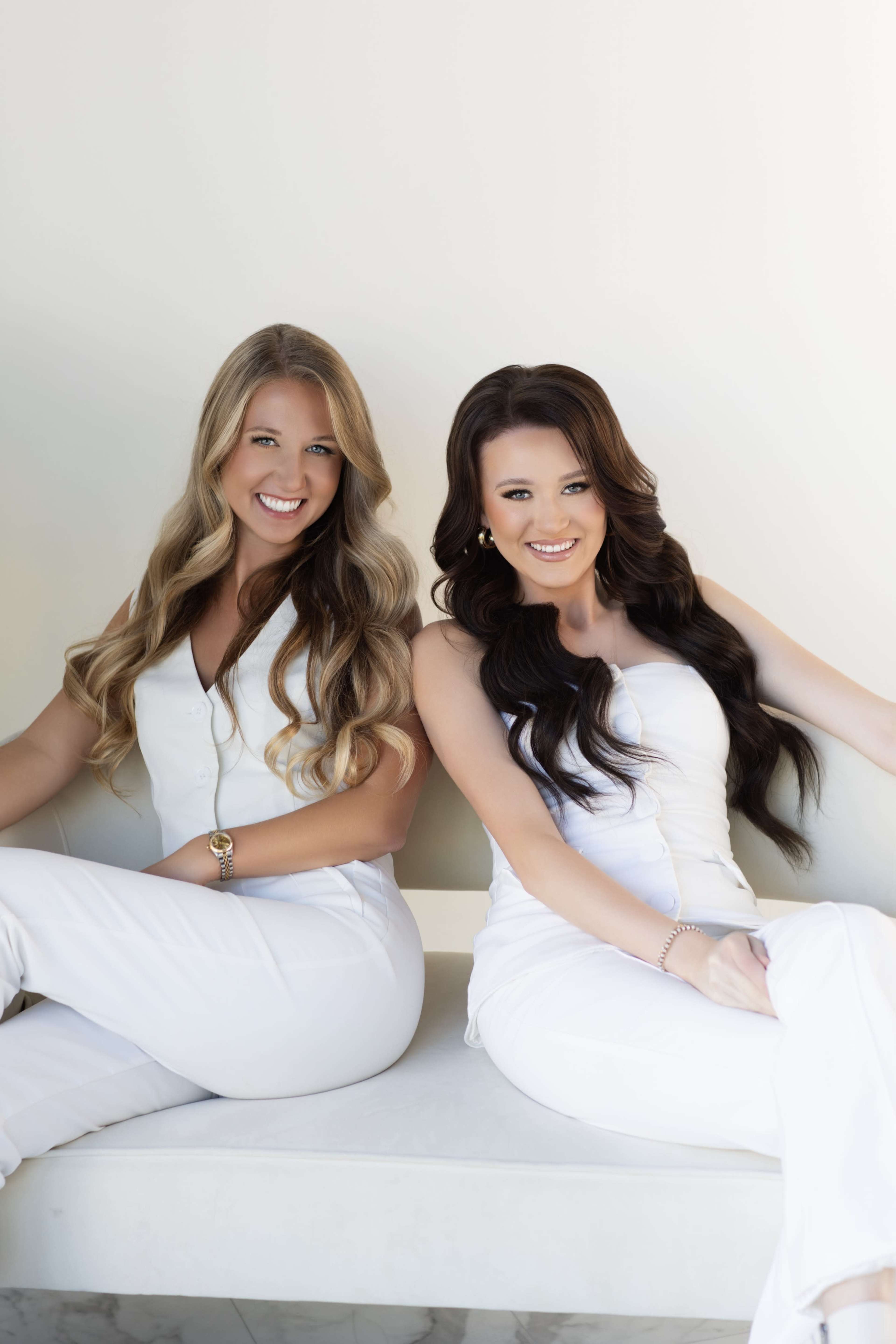 Two women dressed in white outfits are sitting on a light-colored bench with smiles on their faces.