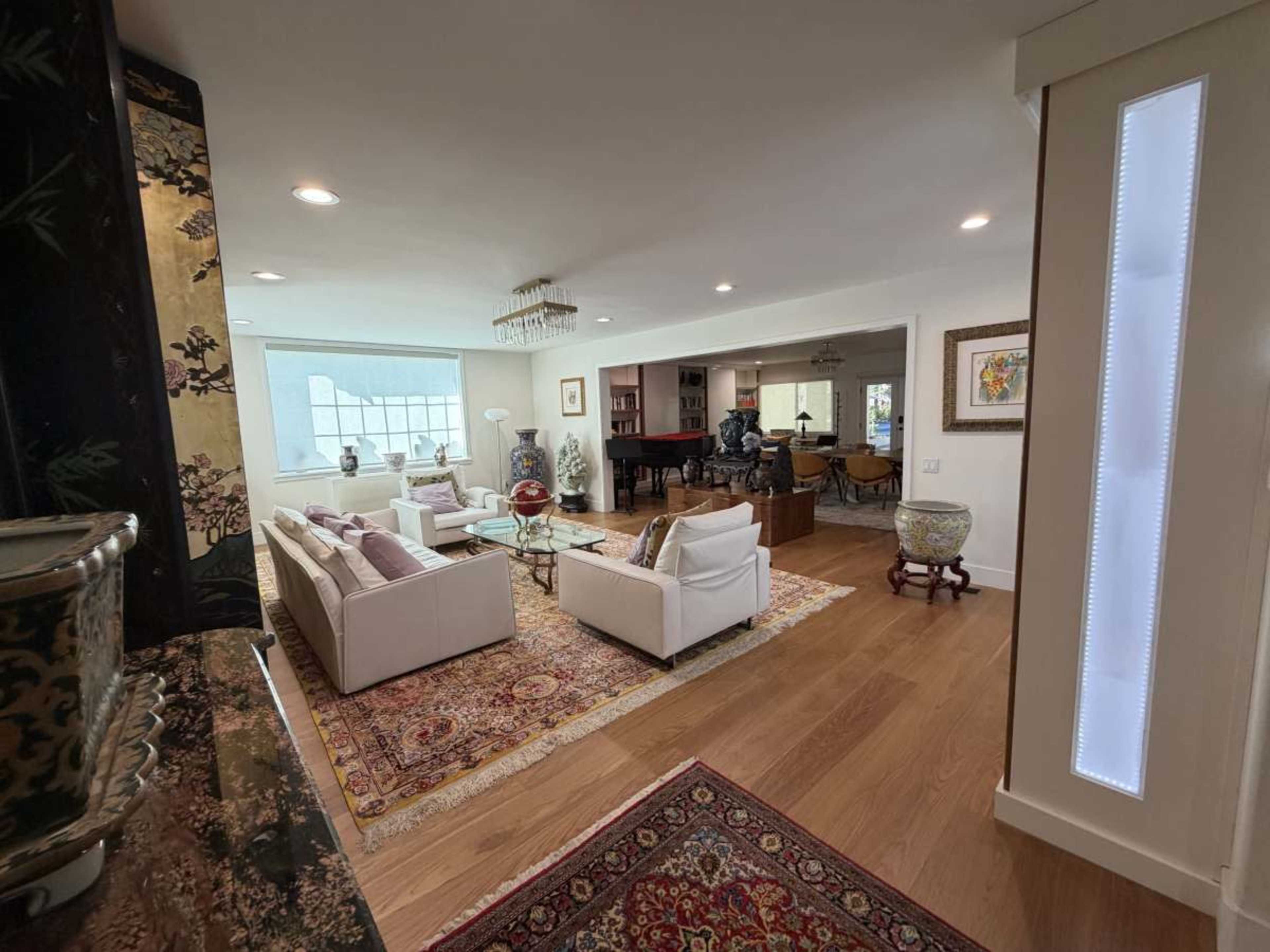 The image shows a spacious living room with white sofas, a coffee table, and a large window, leading to an open area with additional seating and a dark wood bar.