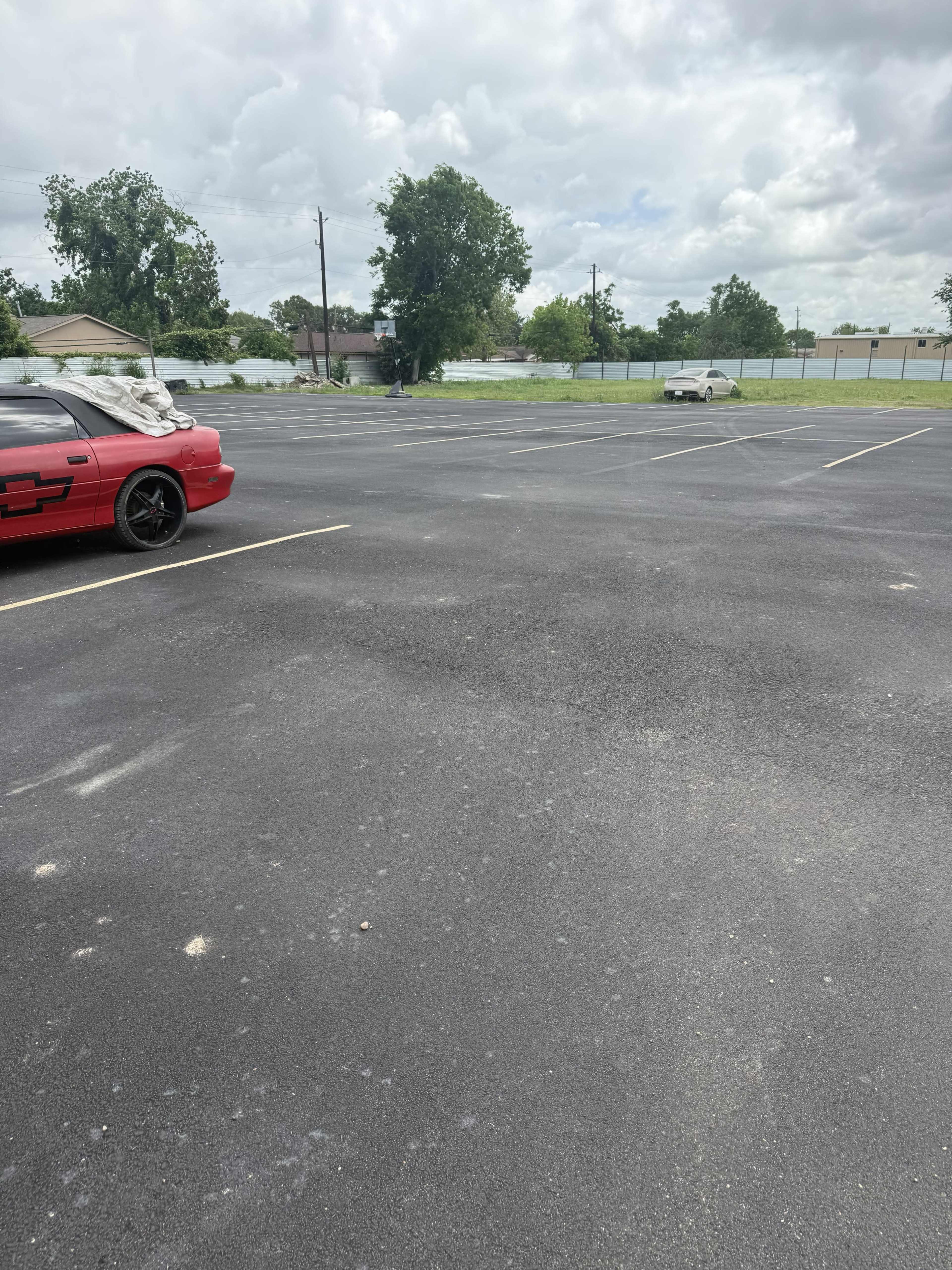The image shows an empty parking lot with a red car parked on one side and a white car in the distance, under a cloudy sky.