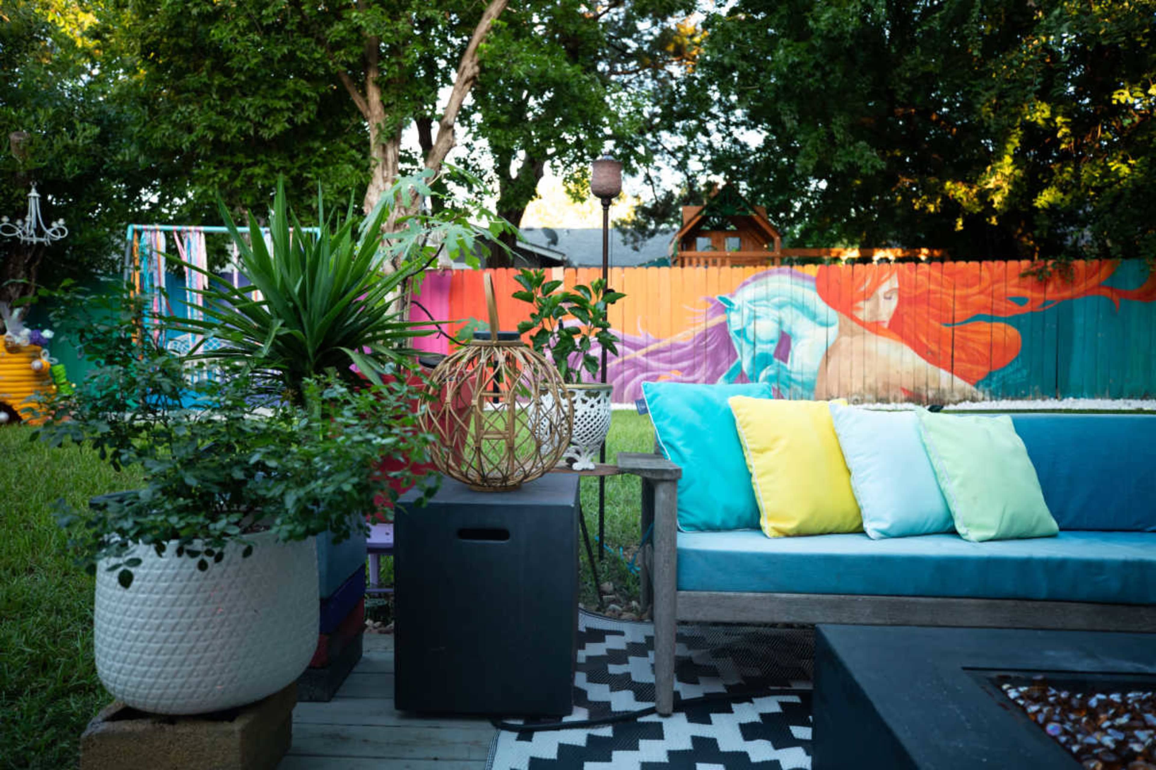 A colorful outdoor patio features a blue sofa with bright cushions, a modern fire pit table, and a vibrant mural on the fence behind it.