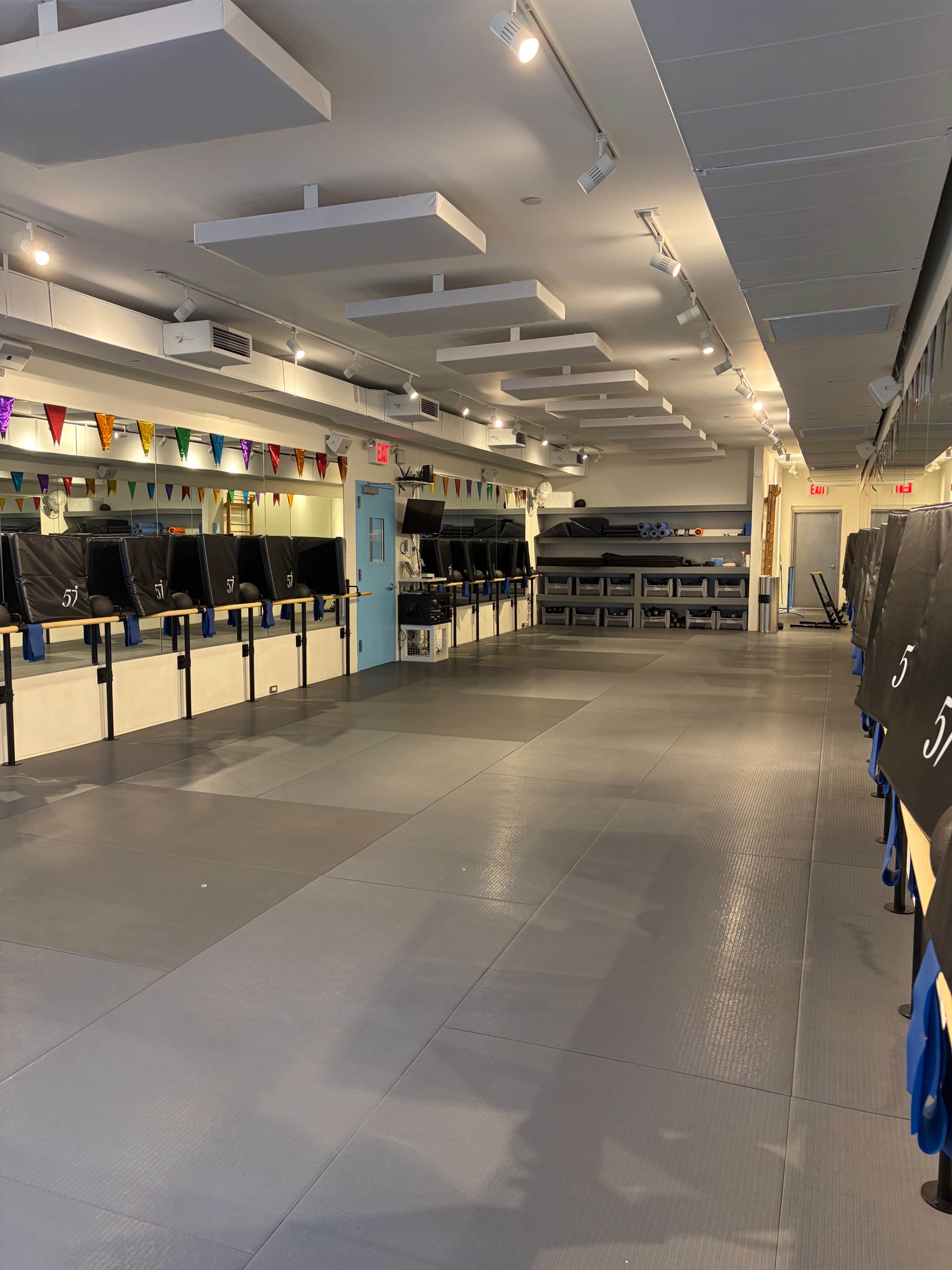 The image shows an empty training studio with mats, mirrors, and colorful buntings hanging from the ceiling.