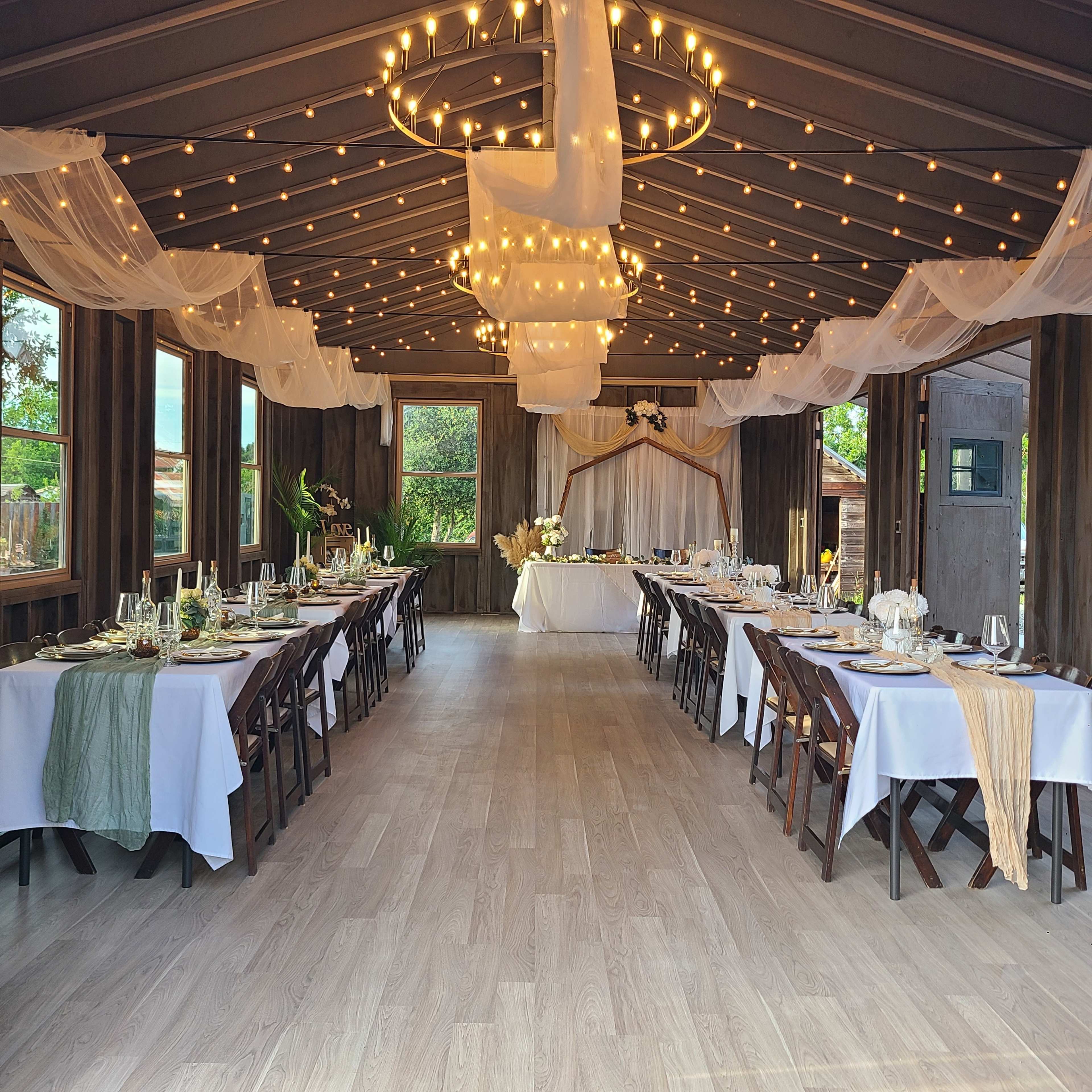 The image shows a decorated event space with two long dining tables set with white tablecloths, elegant centerpieces, and draped fabric overhead, illuminated by string lights.