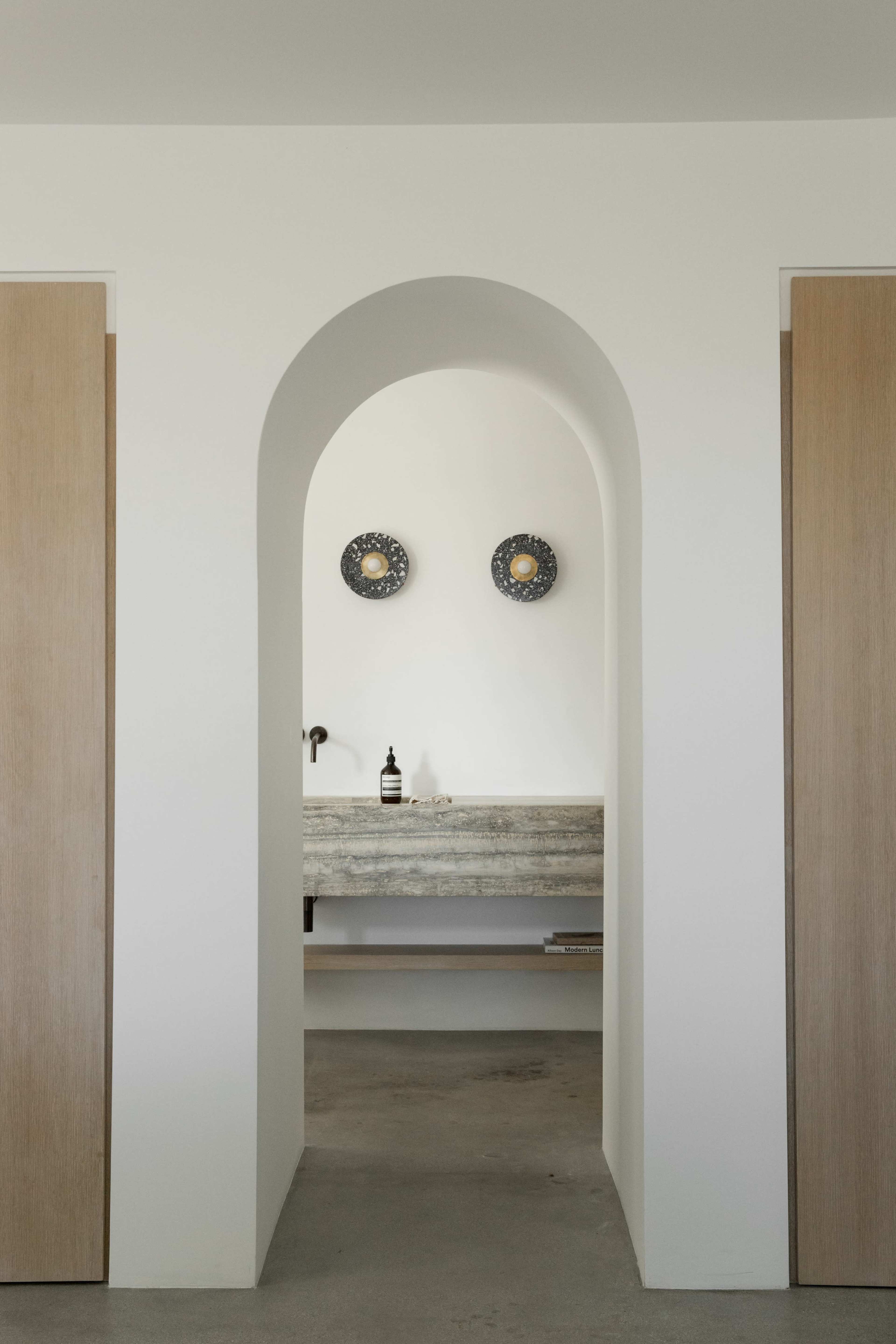 The image features an archway leading into a minimalist room with a stone counter, two decorative wall disks, and wooden doors on either side.