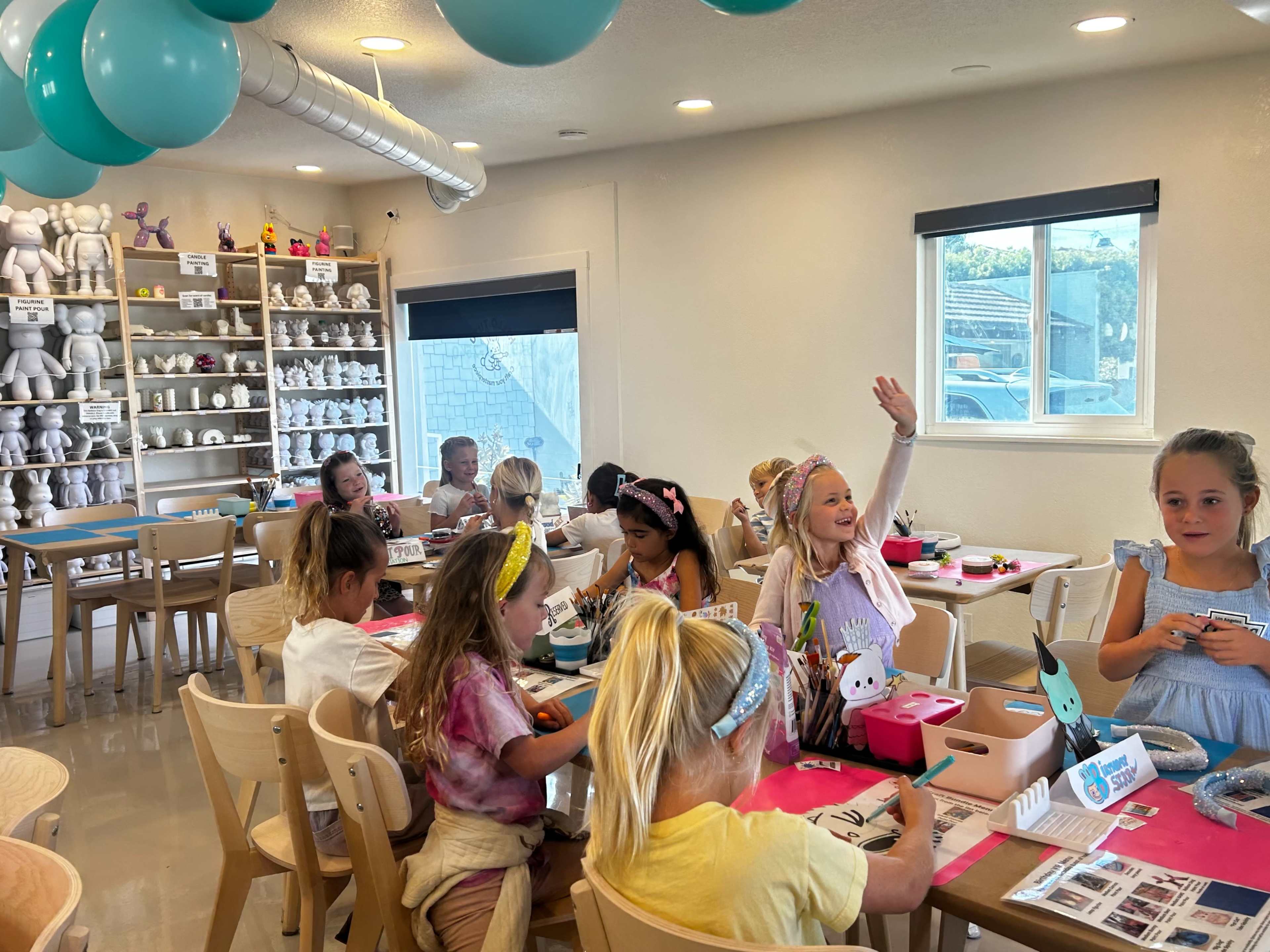 A group of children is sitting at tables in a brightly lit room, engaged in a craft activity with colorful supplies and decorations.