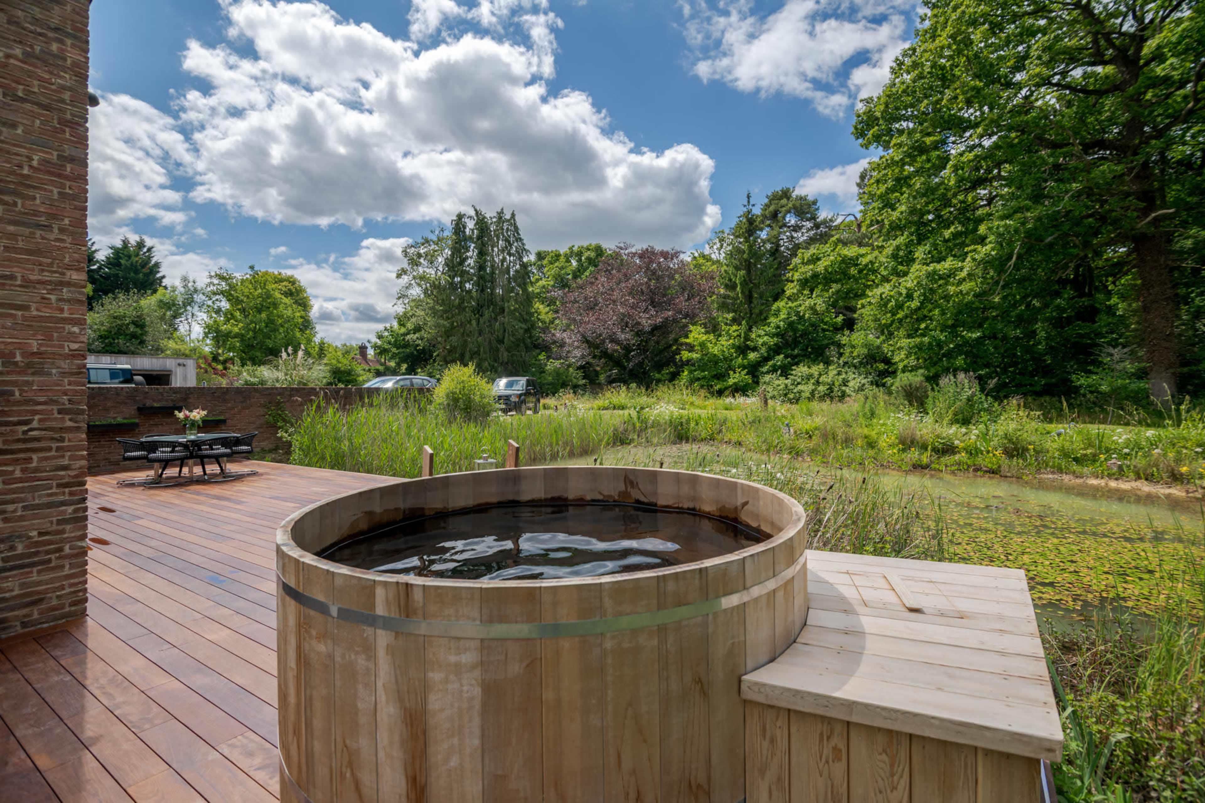 A wooden hot tub sits on a deck overlooking a lush, green landscape with trees and a pond.