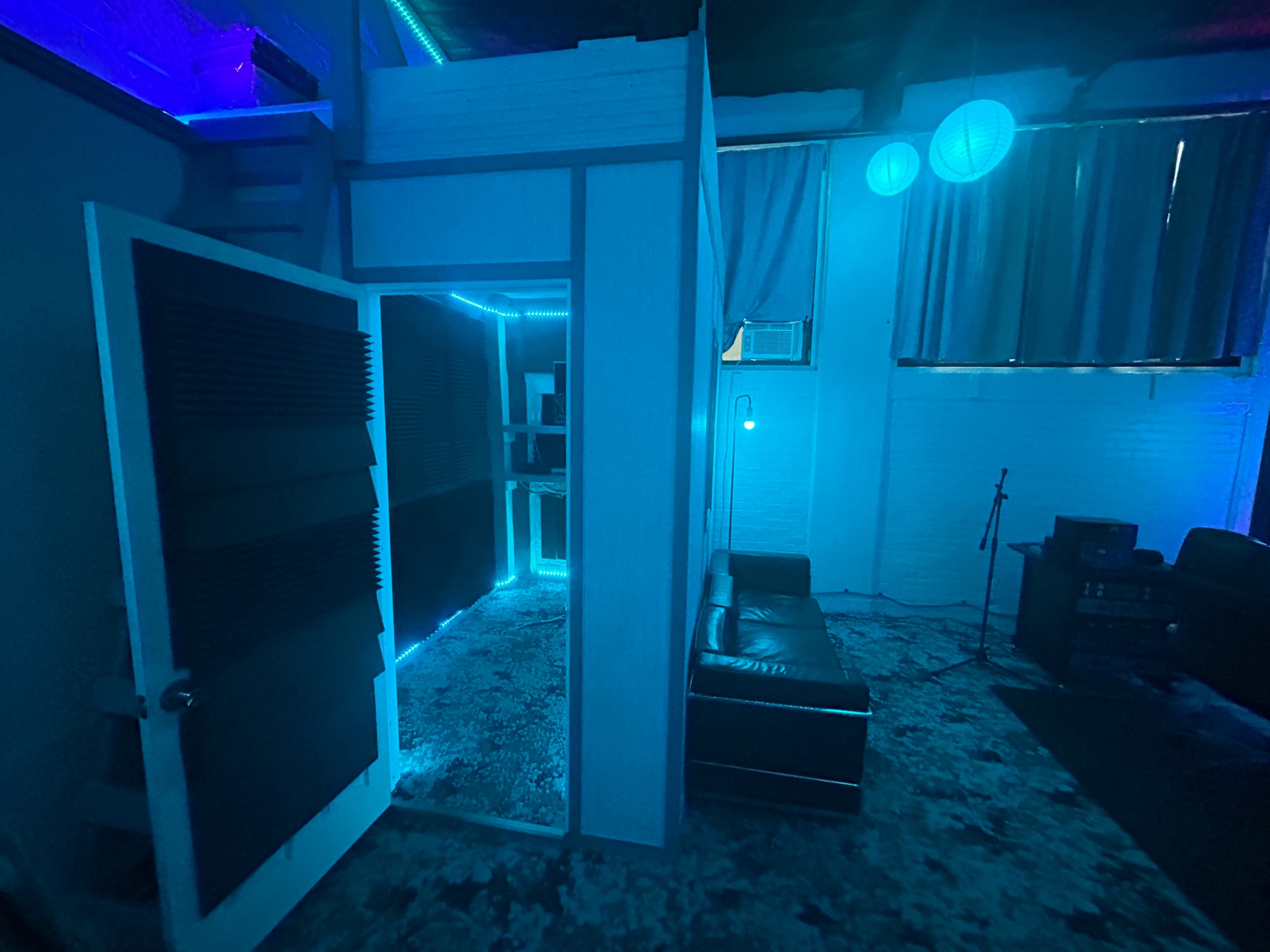 The image shows a dimly lit room with blue neon lights illuminating a couch, a small recording area, and soundproof paneling on the door.