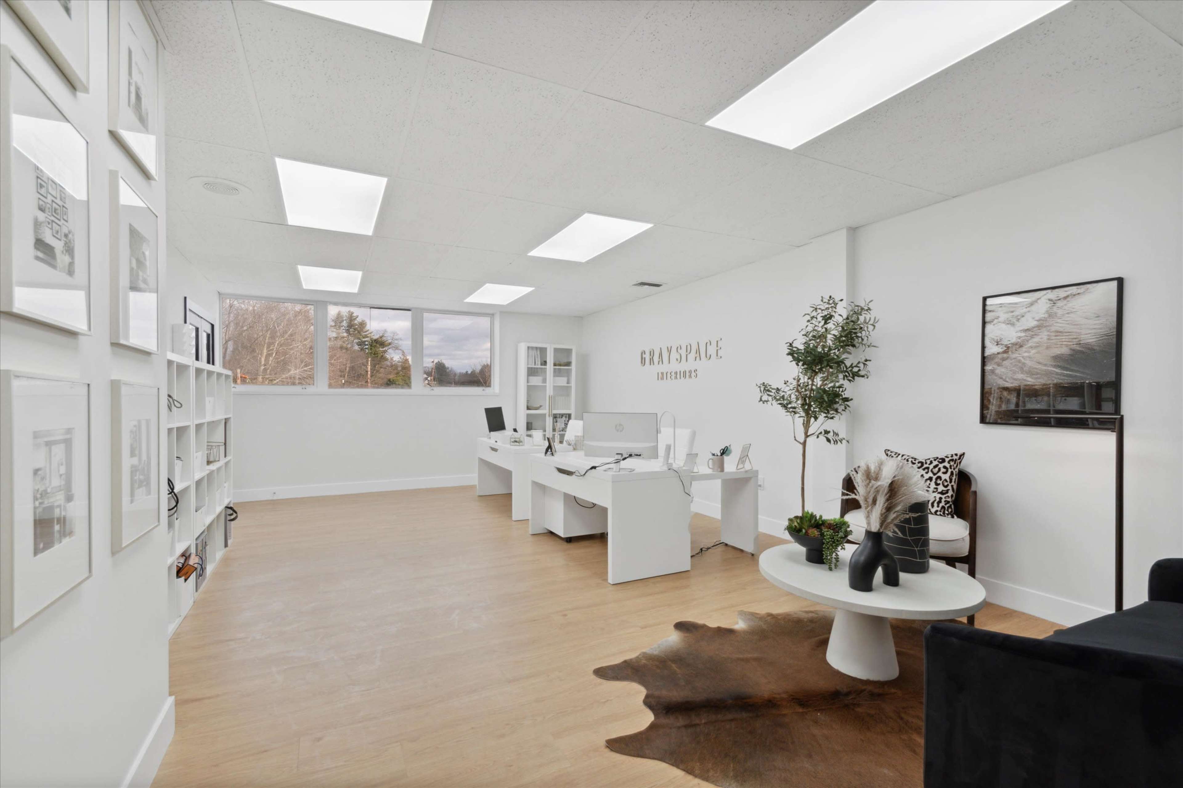 The image shows a modern office space with white walls, wooden floors, and a desk setup, featuring art on one side and a seating area with a plant and decorative items.