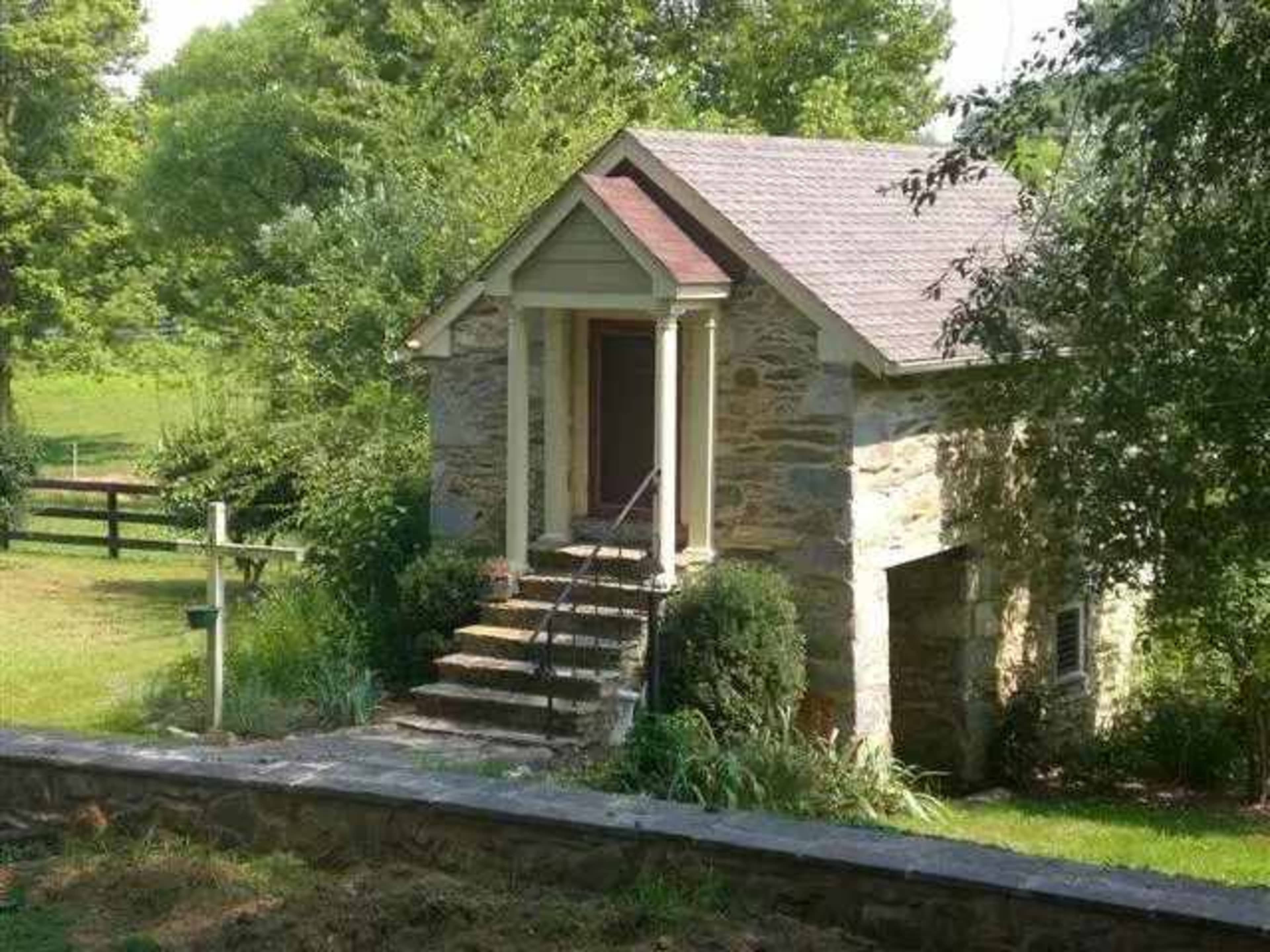 The image shows a small, stone cottage with a gabled roof and a set of steps leading to the front door, surrounded by greenery and trees.