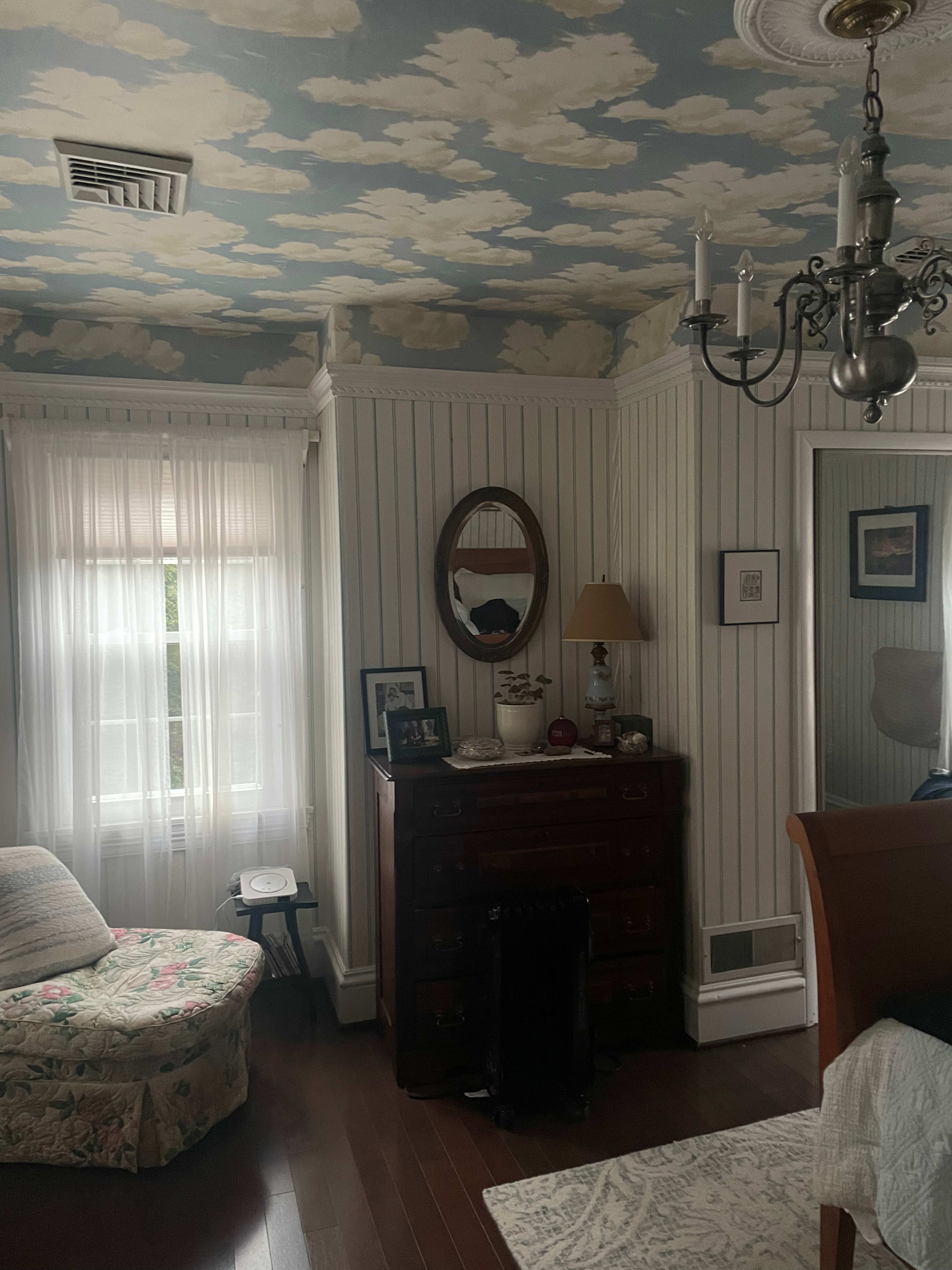 The image depicts a cozy bedroom featuring a floral-patterned chair, a wooden dresser, soft lighting from a lamp, and a chandelier under a cloud-patterned ceiling.