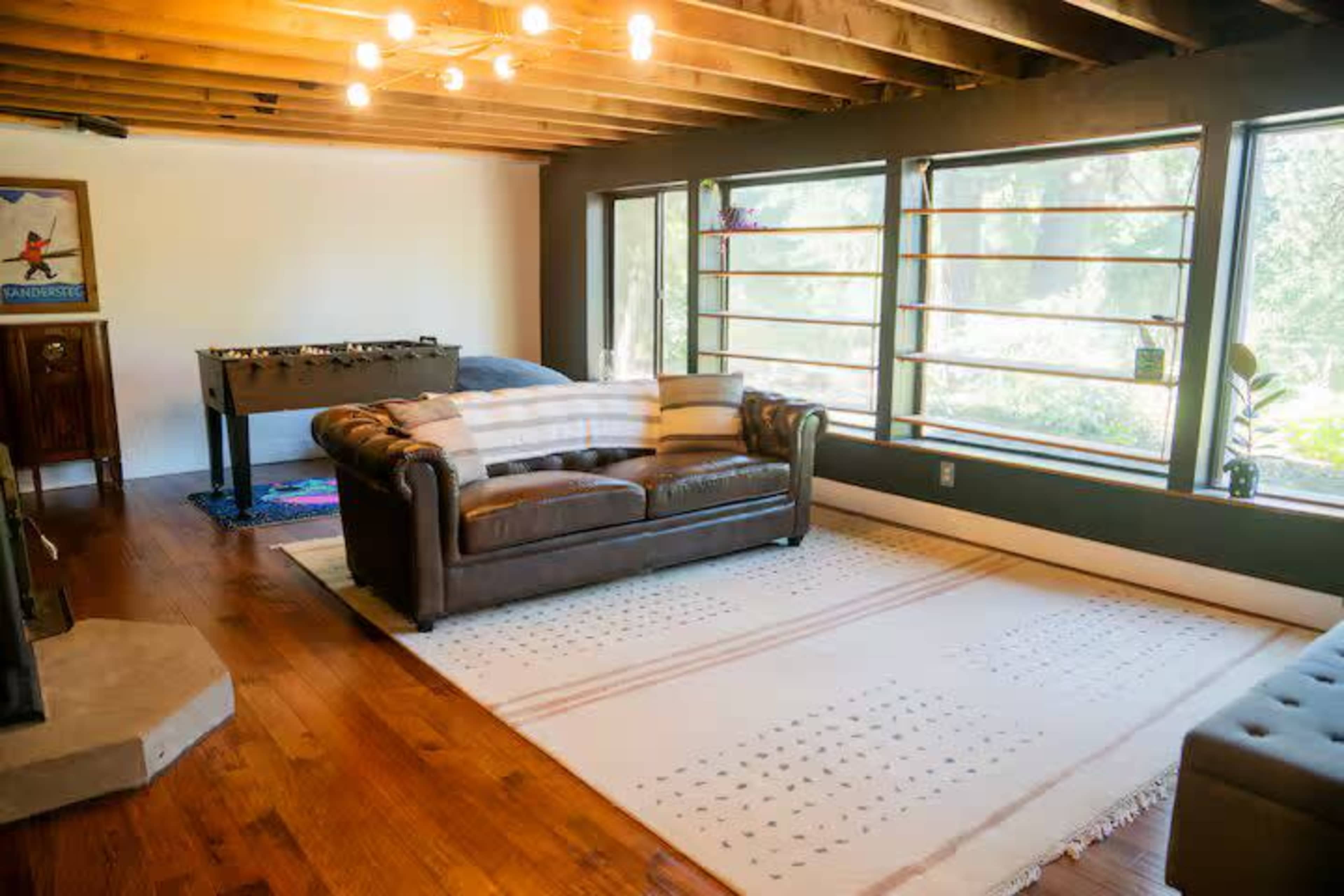 The image shows a spacious living room with a leather sofa, a foosball table, and large windows that let in natural light.