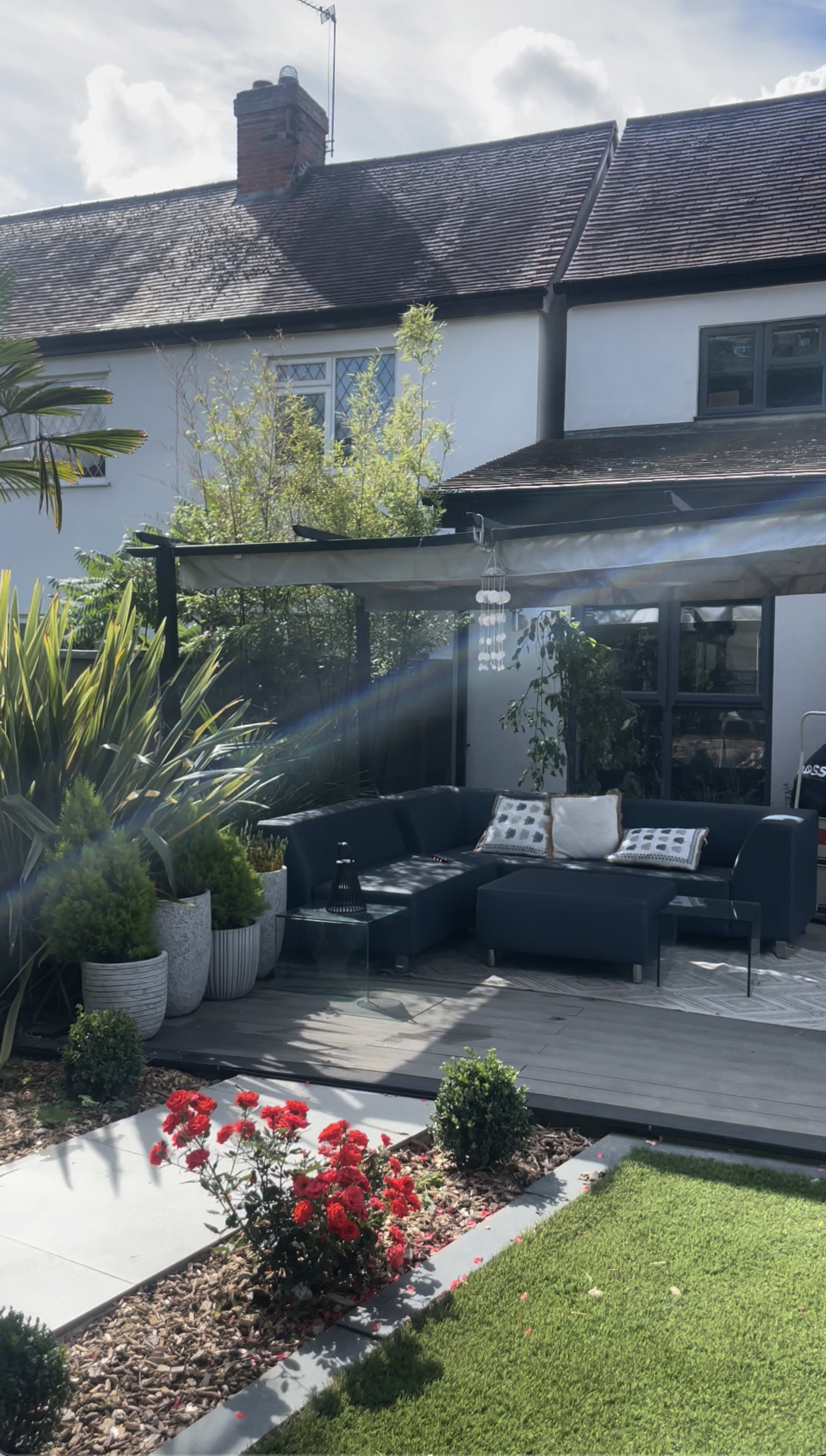 The image shows a landscaped backyard featuring a modern seating area with a pergola, surrounded by plants and a flower garden.