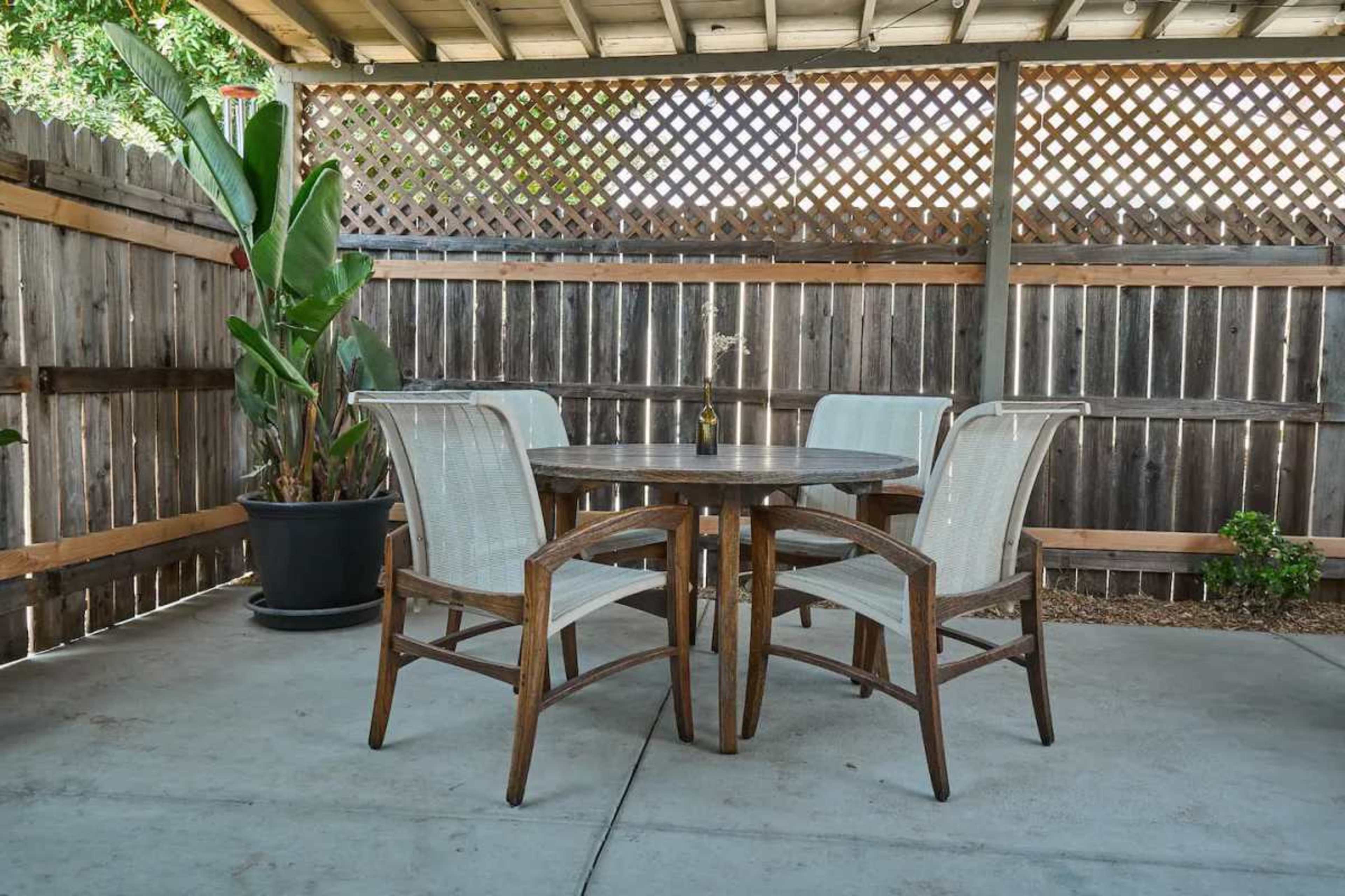 A wooden patio set with four chairs is arranged around a round table beneath a covered area, surrounded by a wooden fence and potted plants.