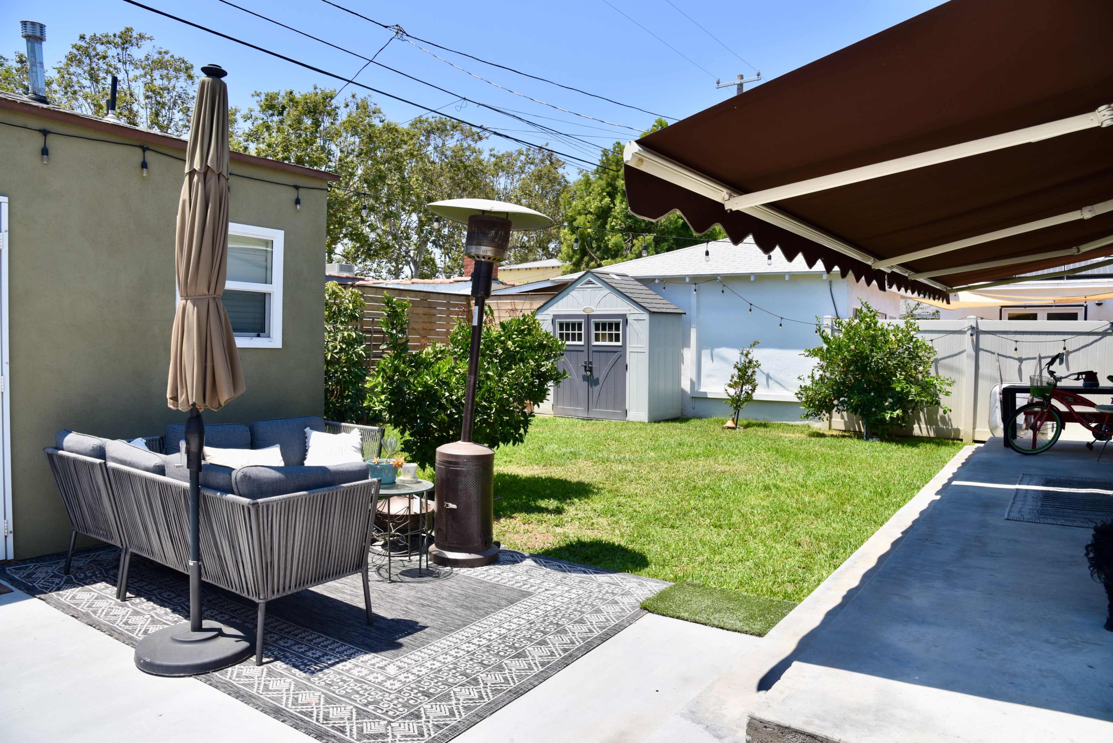 A cozy backyard features a seating area with a patio umbrella, a gas heater, and a small shed surrounded by green grass.