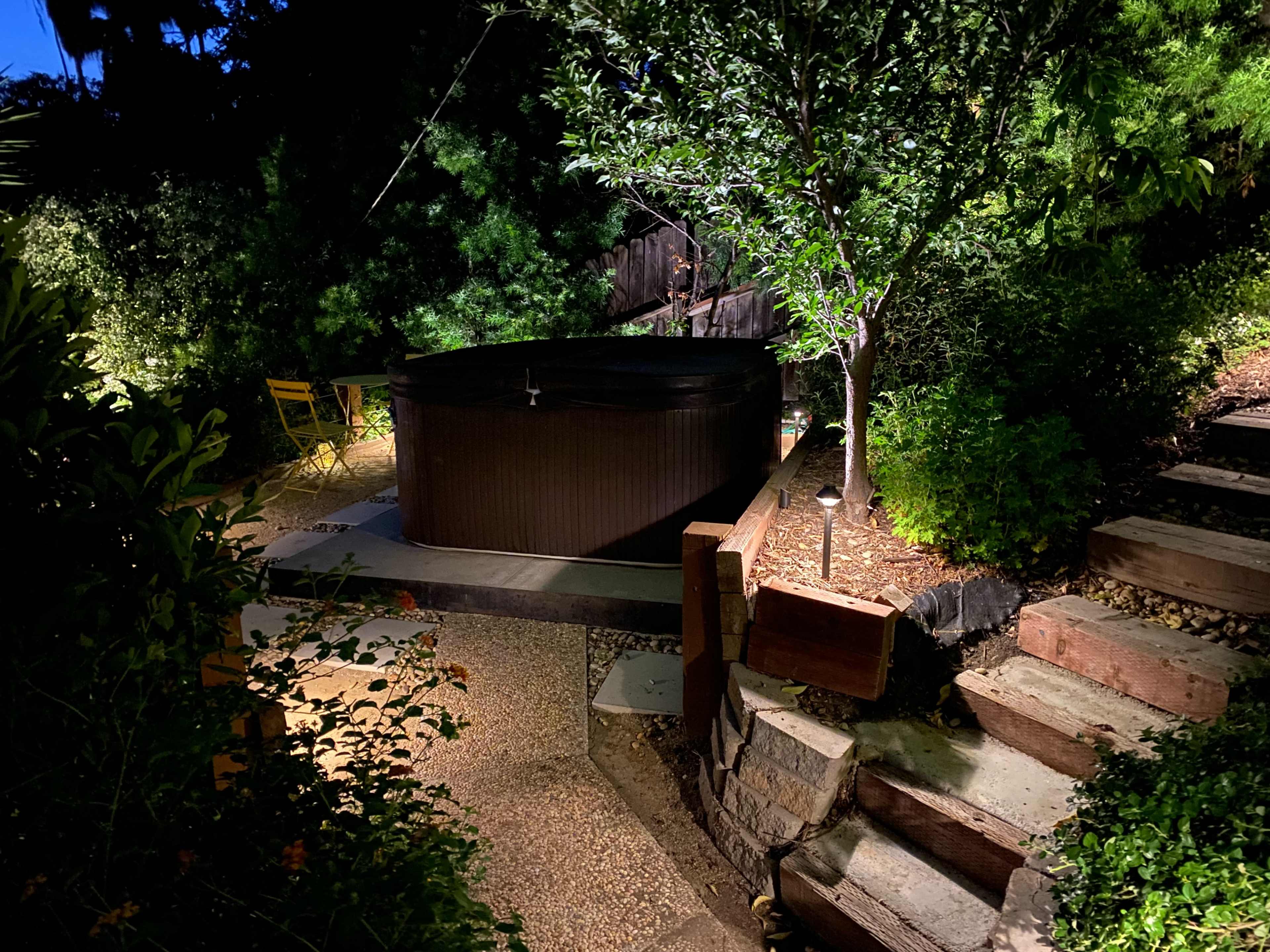 A hot tub is illuminated in a landscaped garden at night, surrounded by trees and wooden steps.