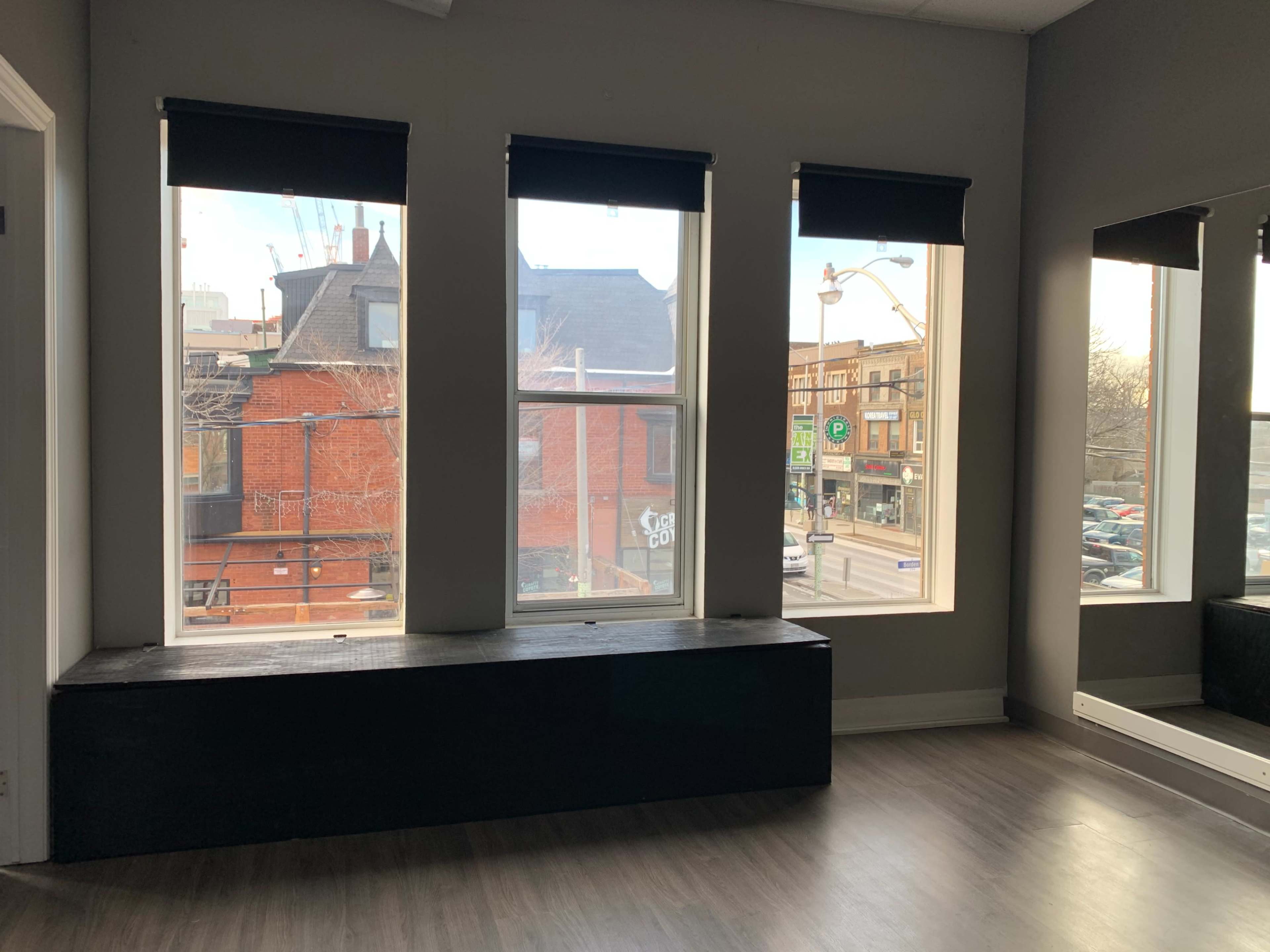 The image shows a room with three large windows, a black bench beneath the windows, and a wall-mounted mirror, overlooking a street with parked cars and buildings outside.