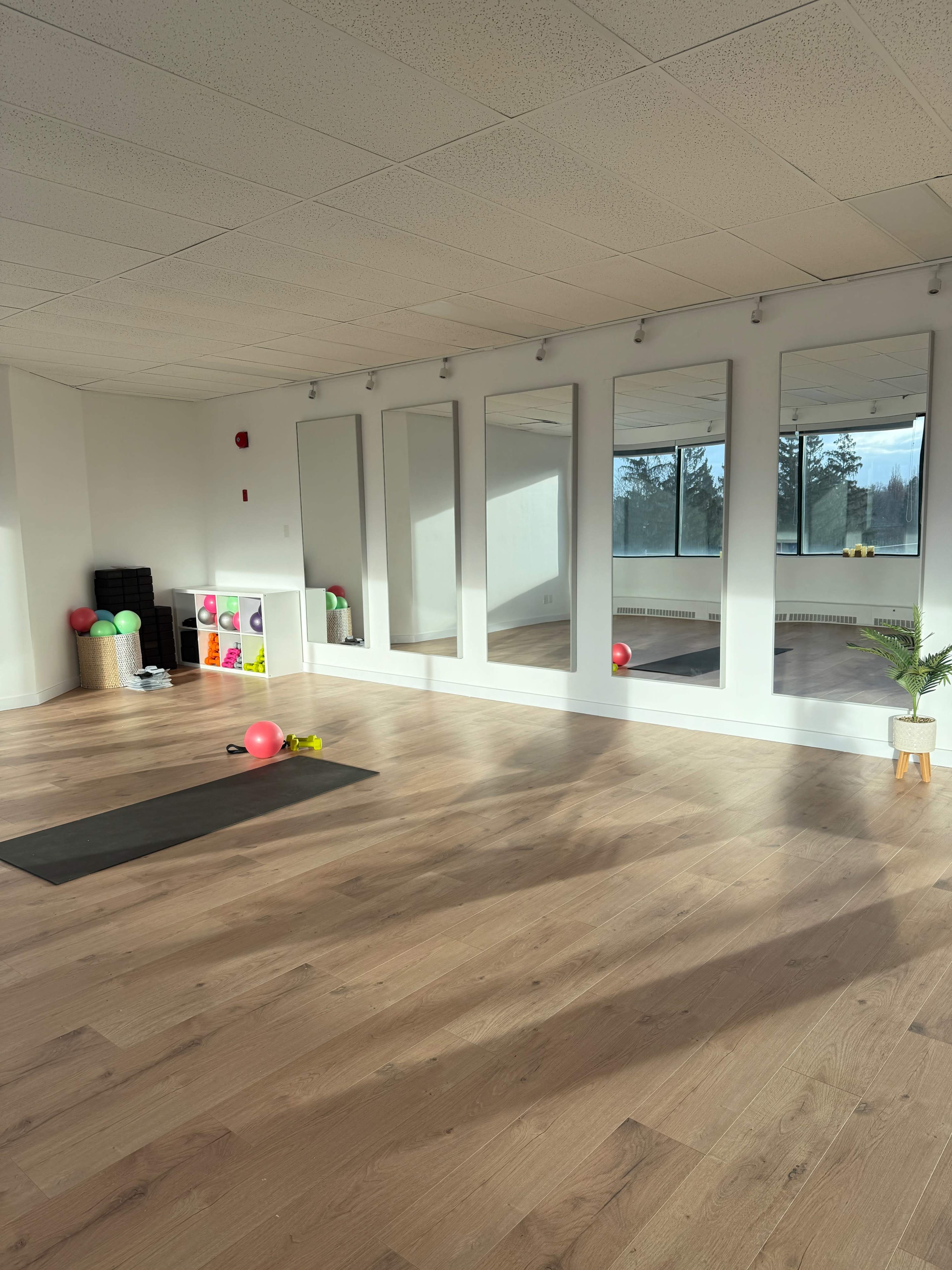 The image shows a bright and spacious exercise studio with wooden floors, mirrors on one wall, and fitness equipment scattered around.