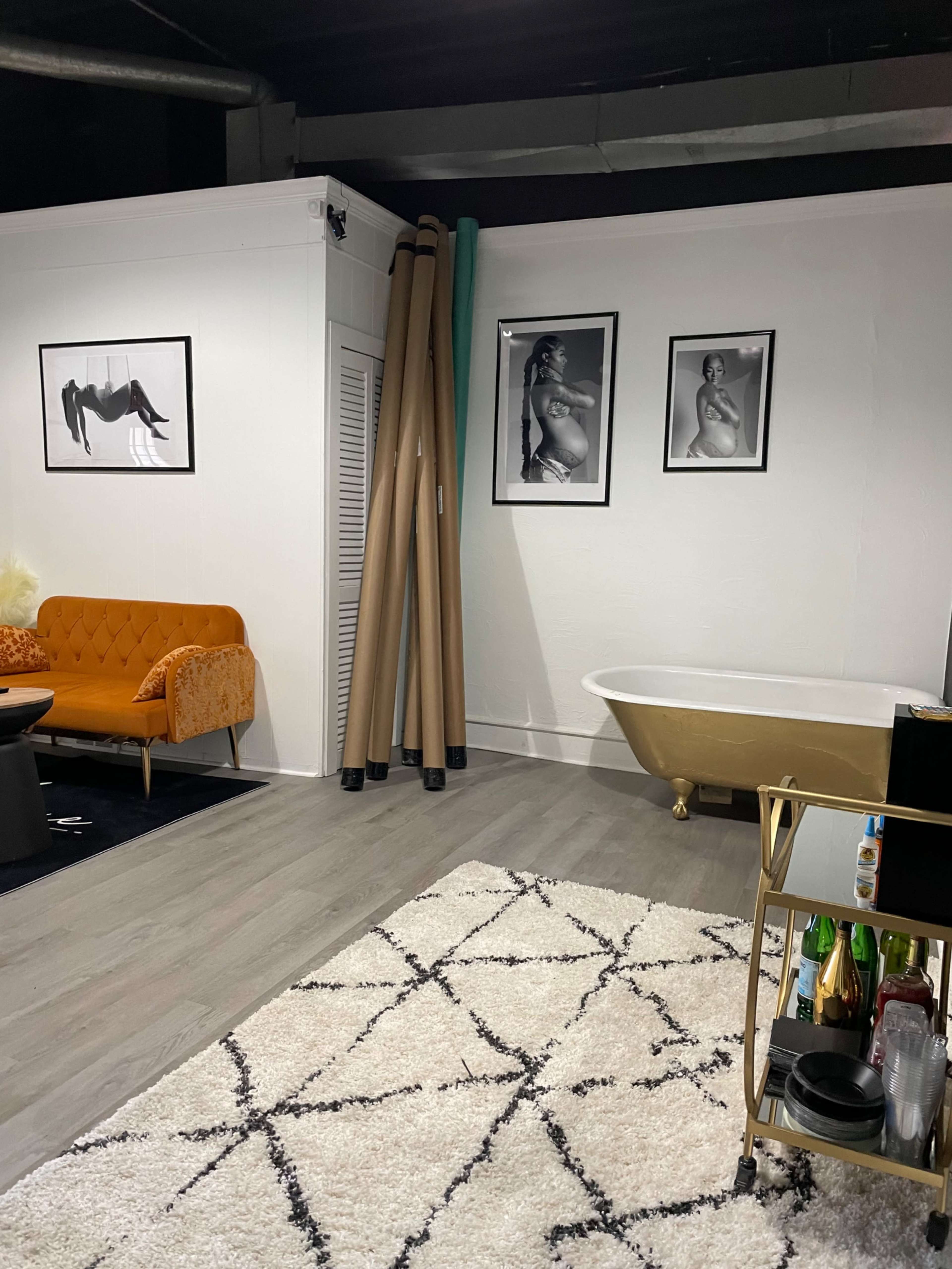 The image shows a room with a vintage bathtub, an orange sofa, a small bar cart with beverages, and several black-and-white photographs on the wall.