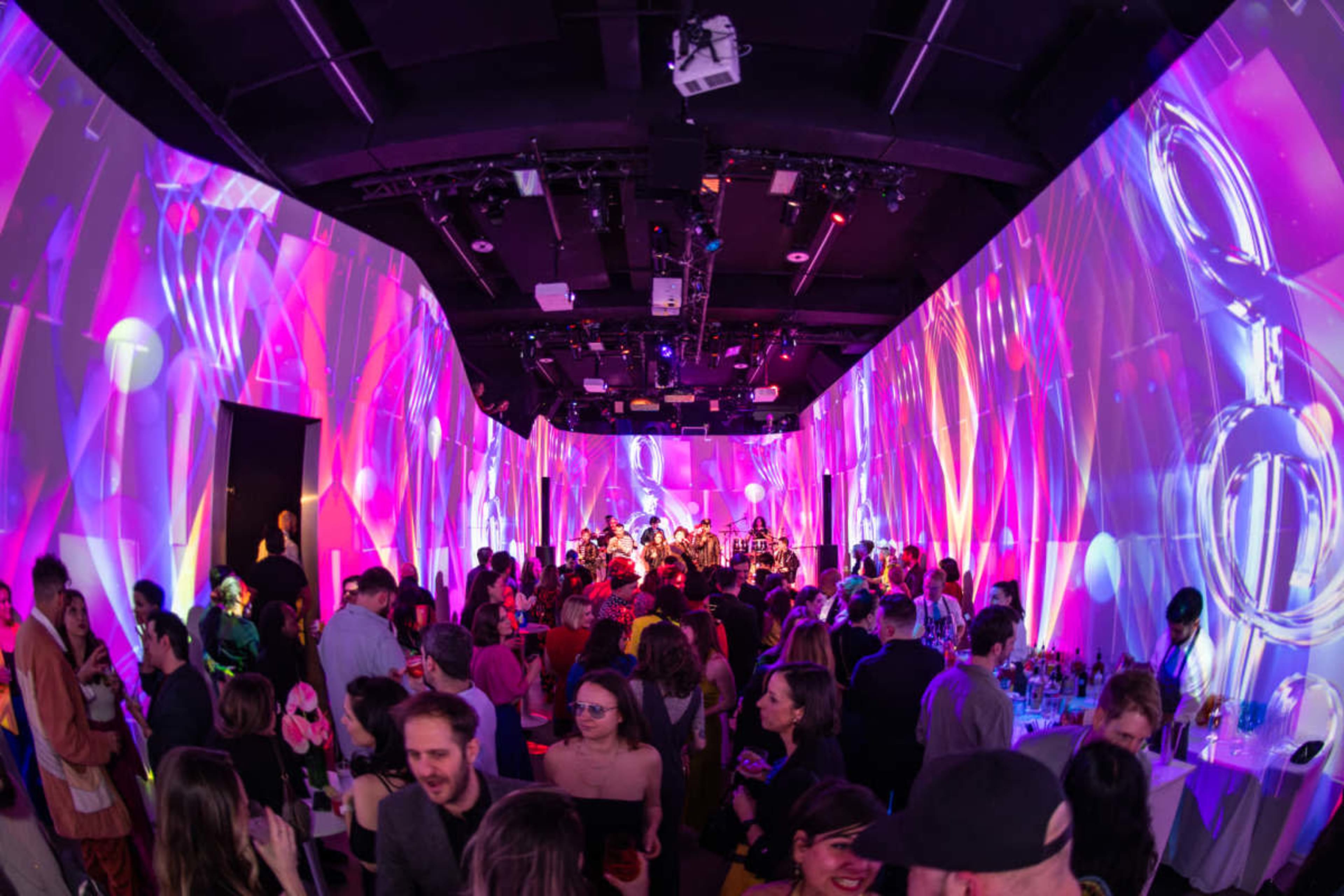 A crowded event space features vibrant projections of colorful patterns on the walls while people mingle and enjoy the atmosphere.