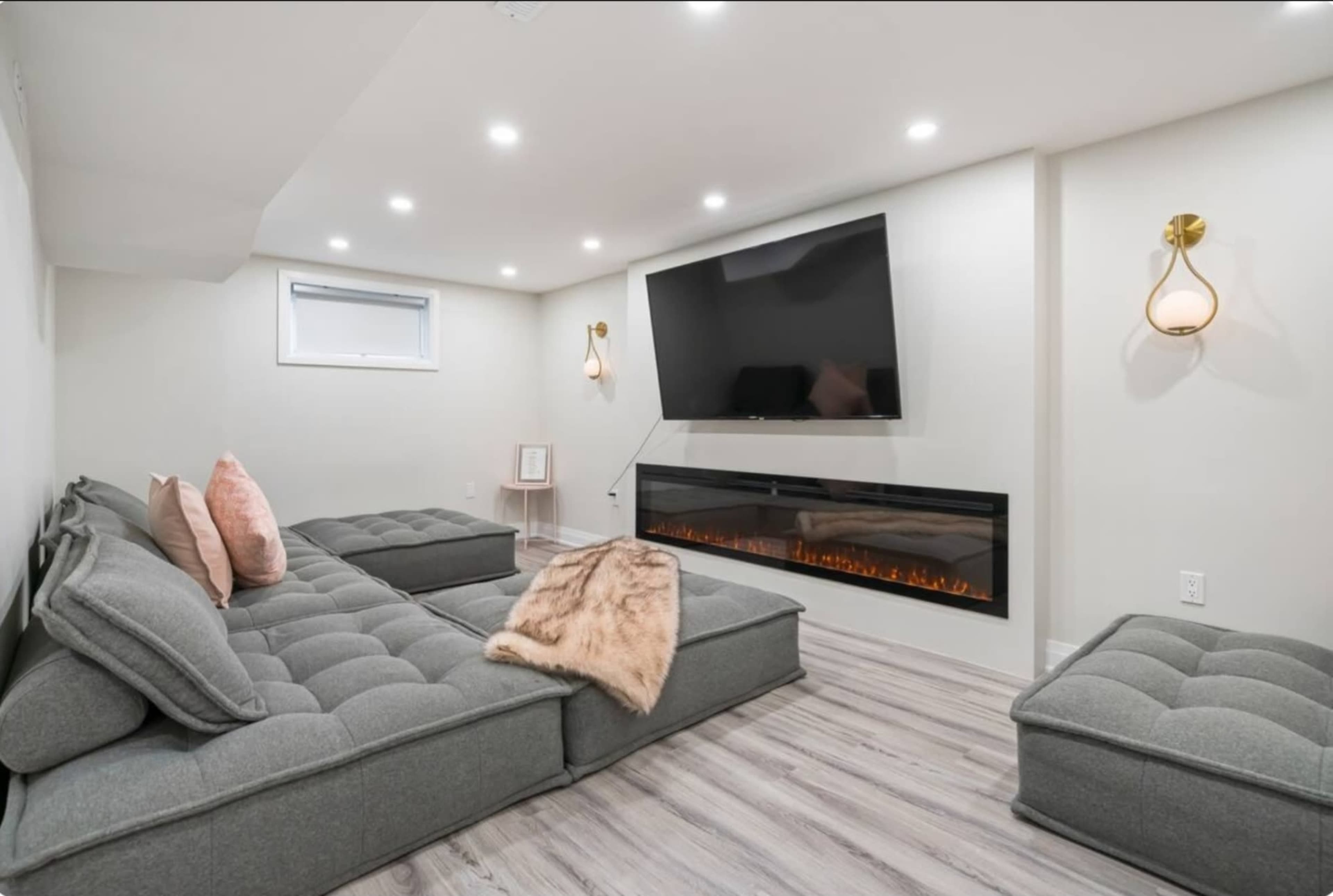 The image shows a modern living room with a gray sectional sofa, a wall-mounted TV, and an electric fireplace beneath.