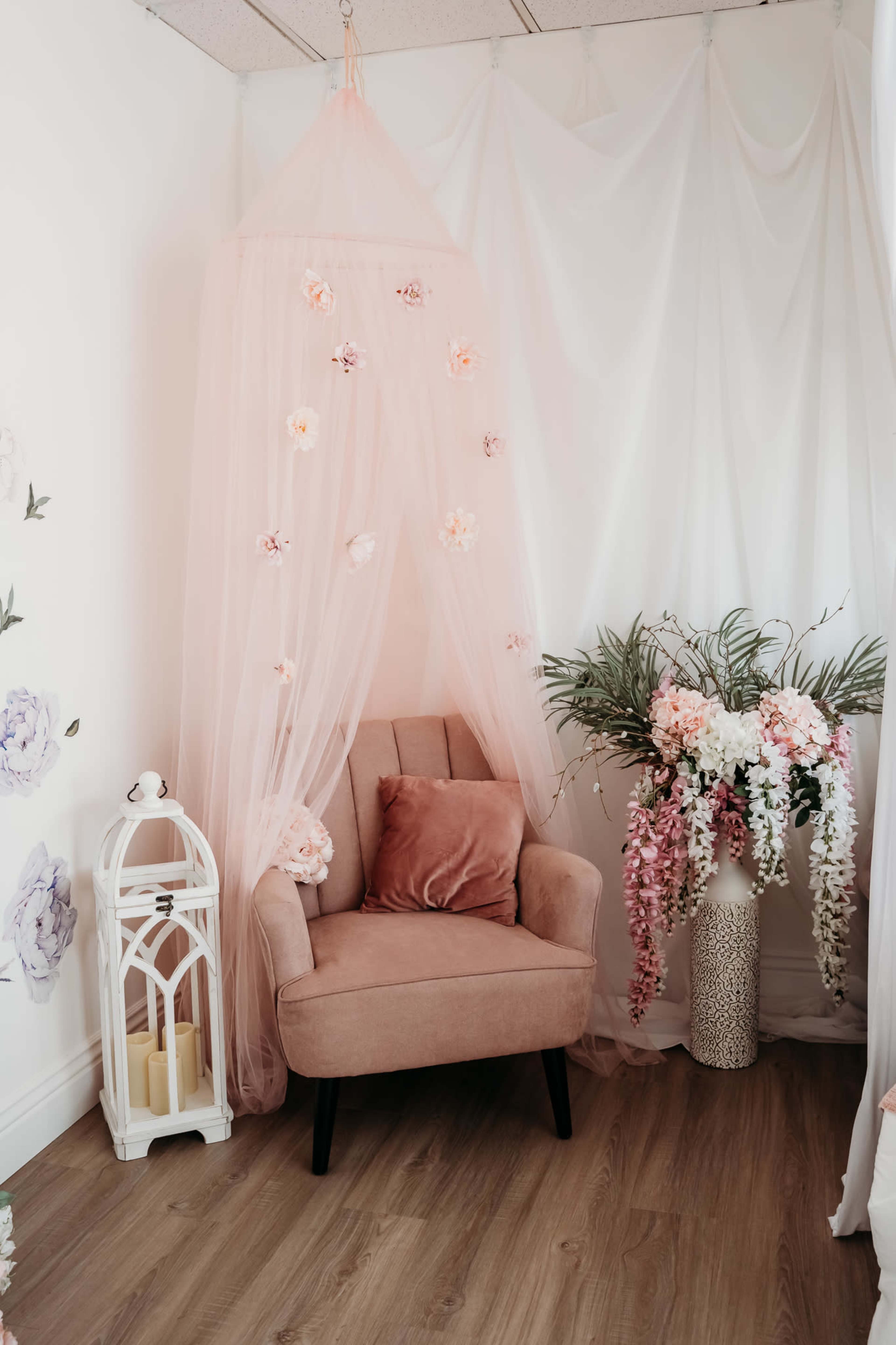 A soft pink armchair is positioned next to a floral arrangement and draped fabric in a cozy corner of a room.