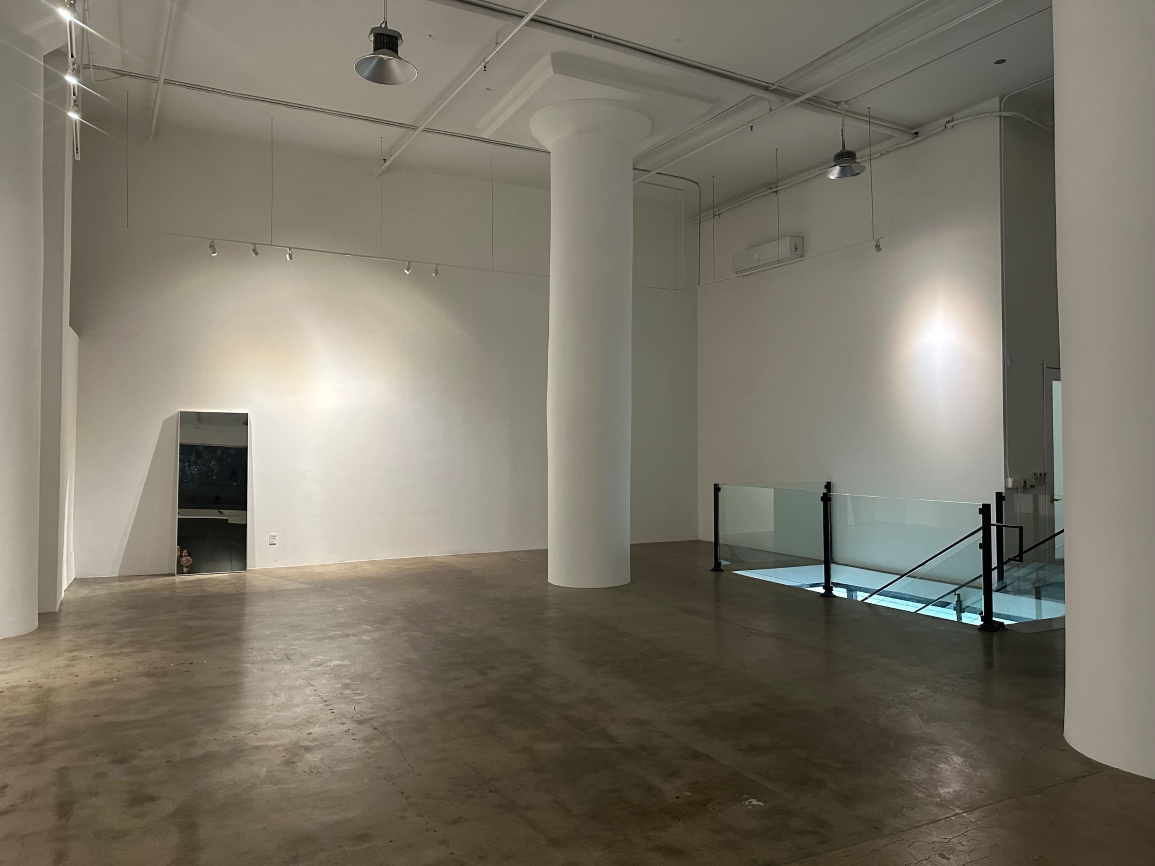 The image shows a spacious, minimally furnished interior with concrete flooring, white walls, and a central support column.