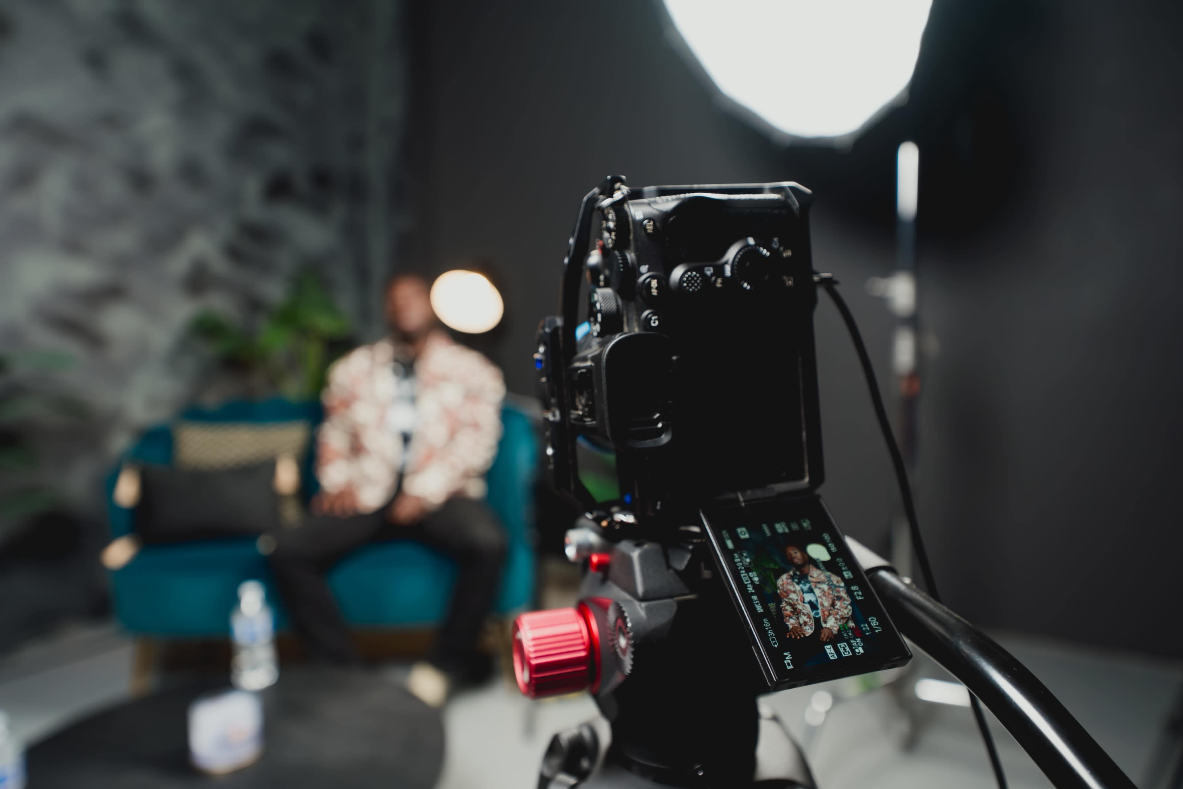A camera on a tripod captures an individual seated on a blue couch in a studio setting.