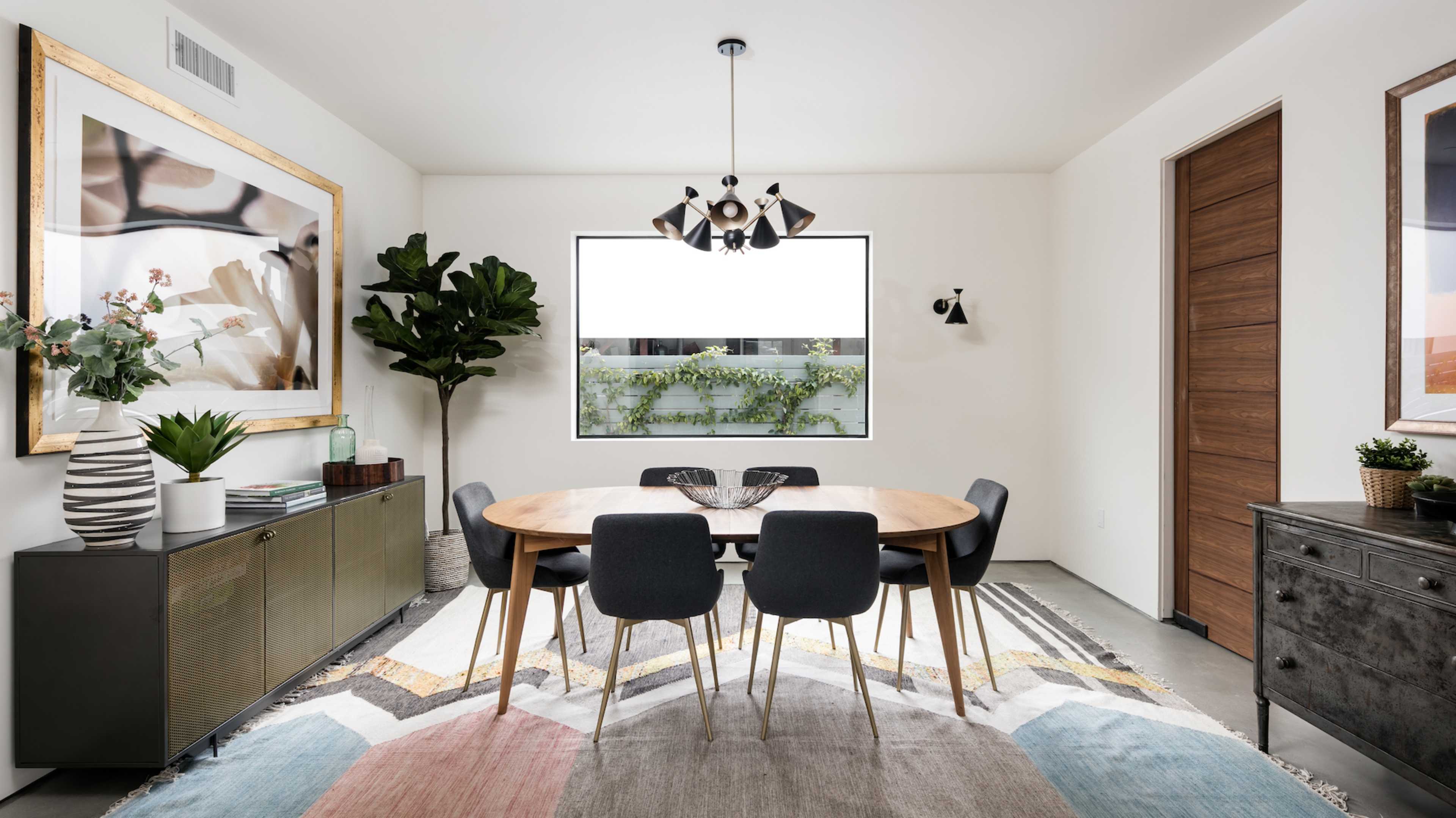 A modern dining room features a round wooden table surrounded by black chairs, with large windows, decorative artwork, and green plants.
