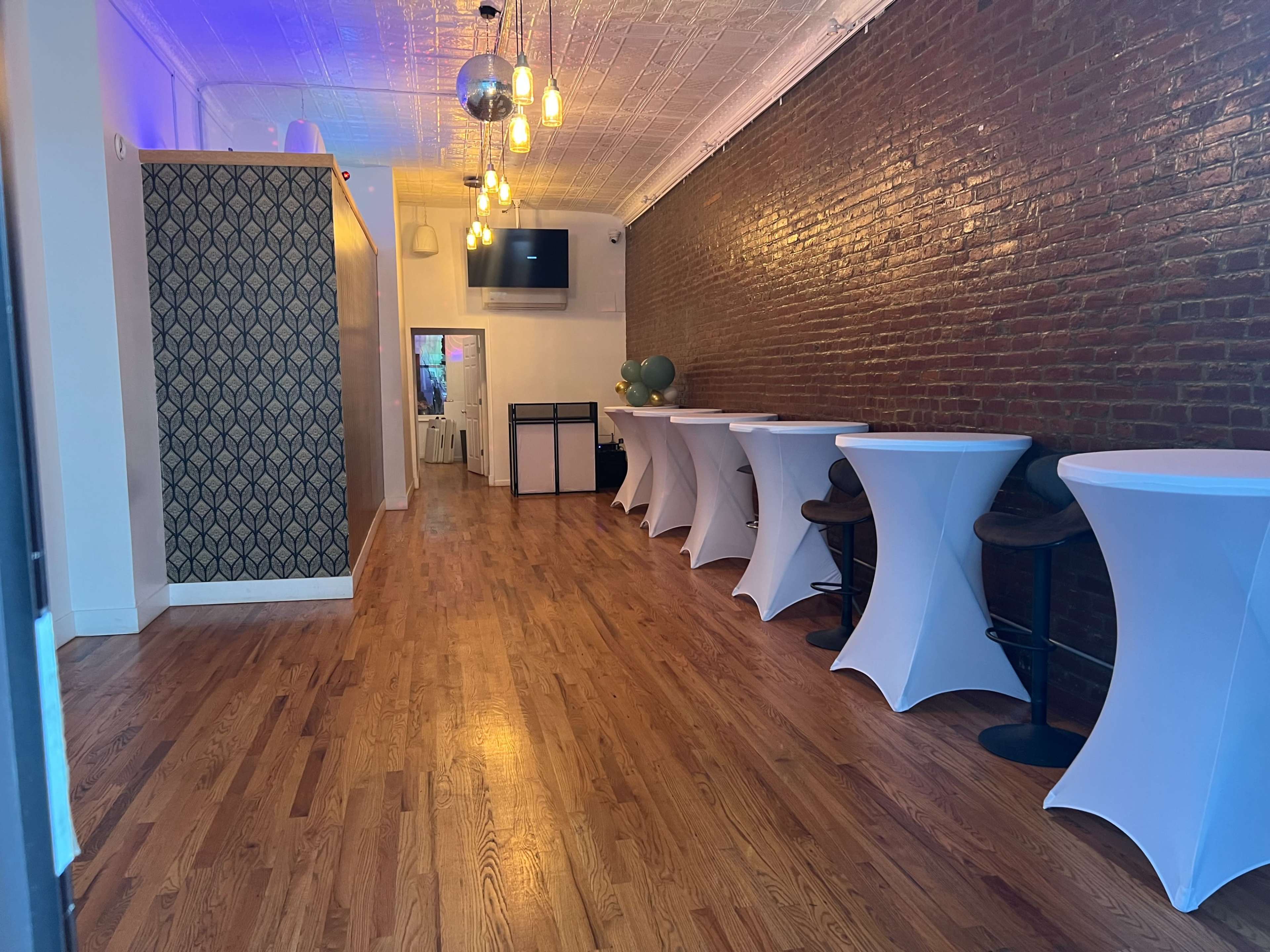 A long, narrow event space with wooden flooring, a brick wall, and several white cocktail tables arranged along one side.