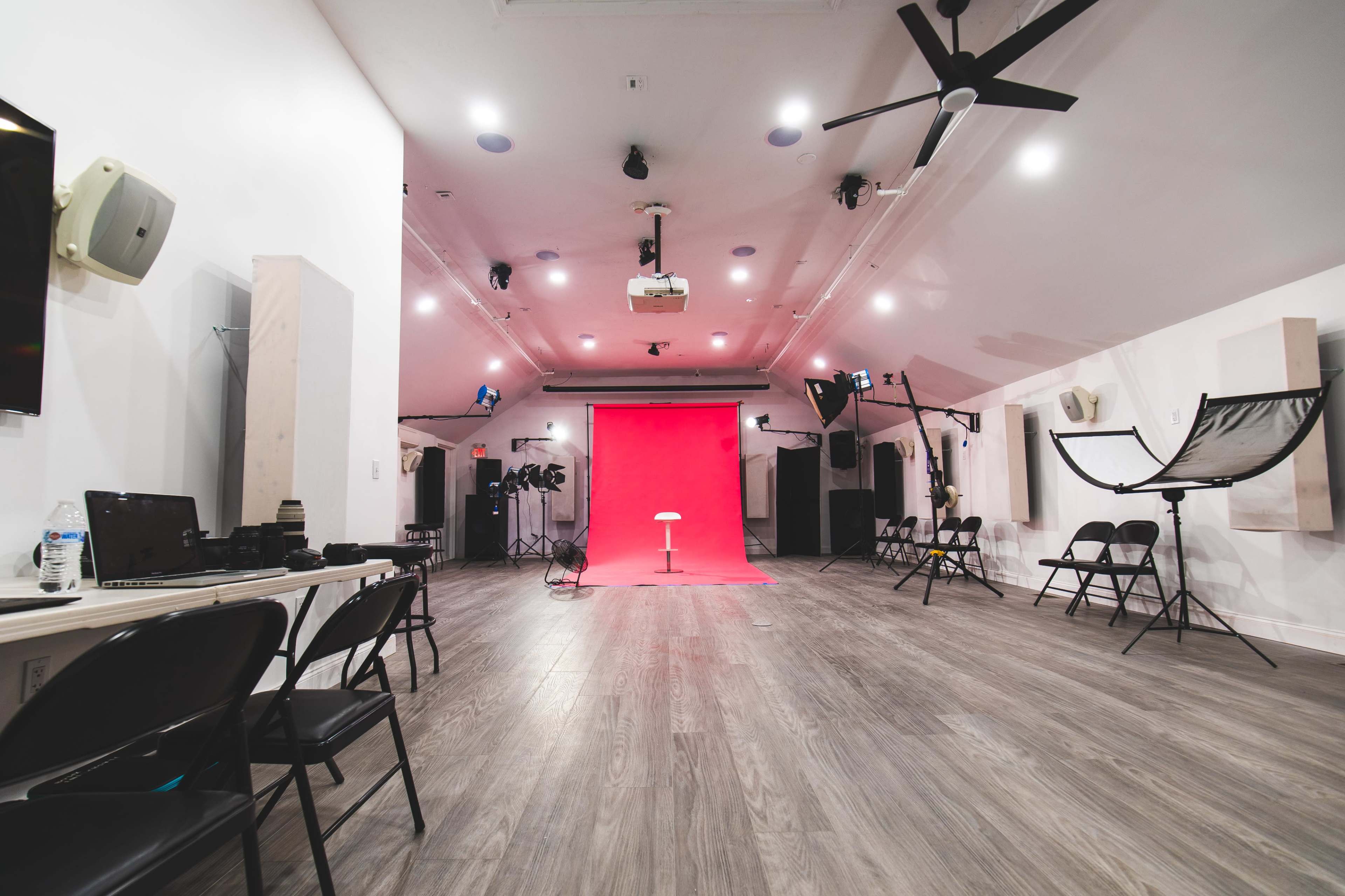The image shows a spacious photography studio with a red backdrop, a white stool in the center, and various lighting equipment and chairs arranged around the room.