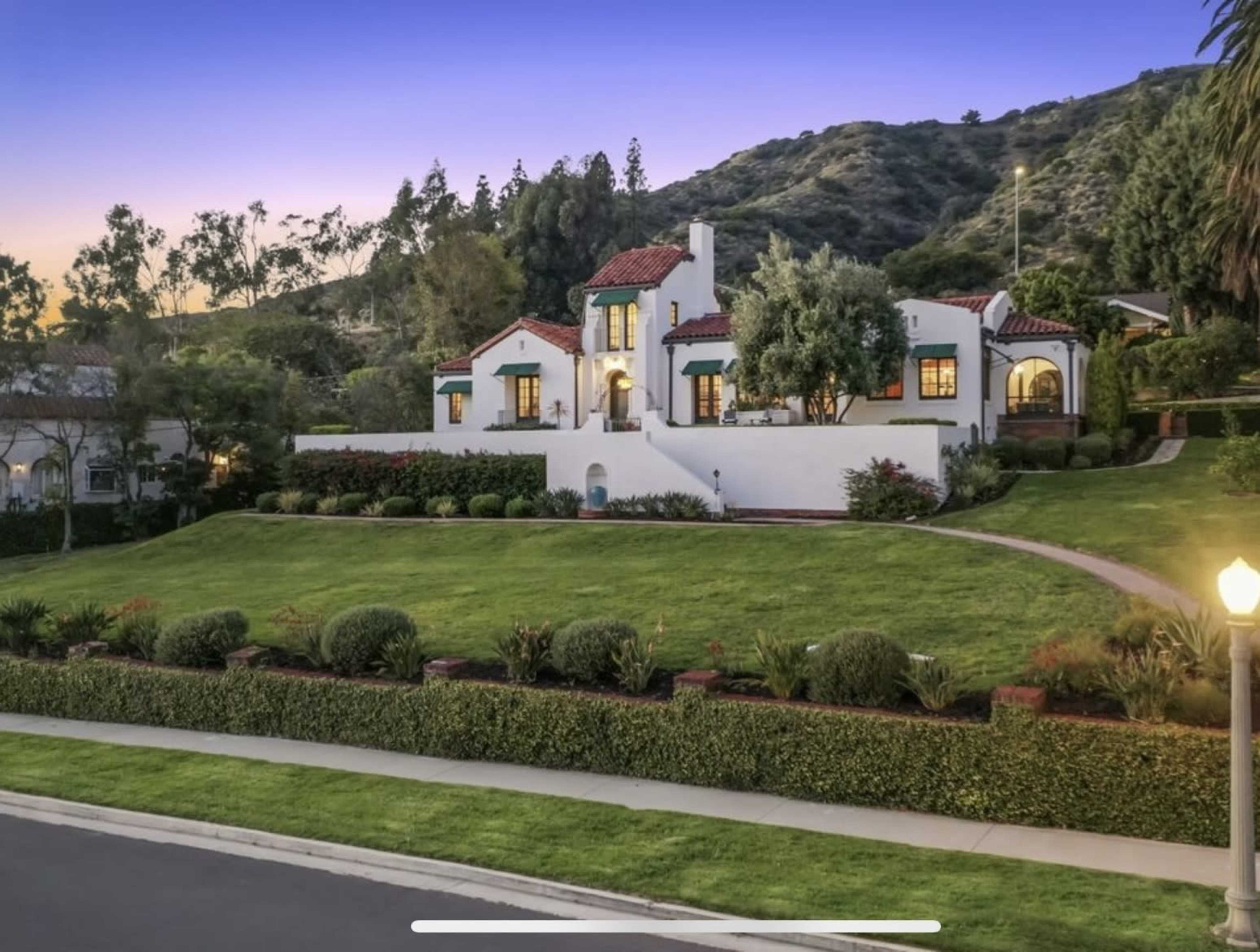 A Mediterranean-style house with red tile roofing sits atop a landscaped hill, surrounded by greenery and pathways.