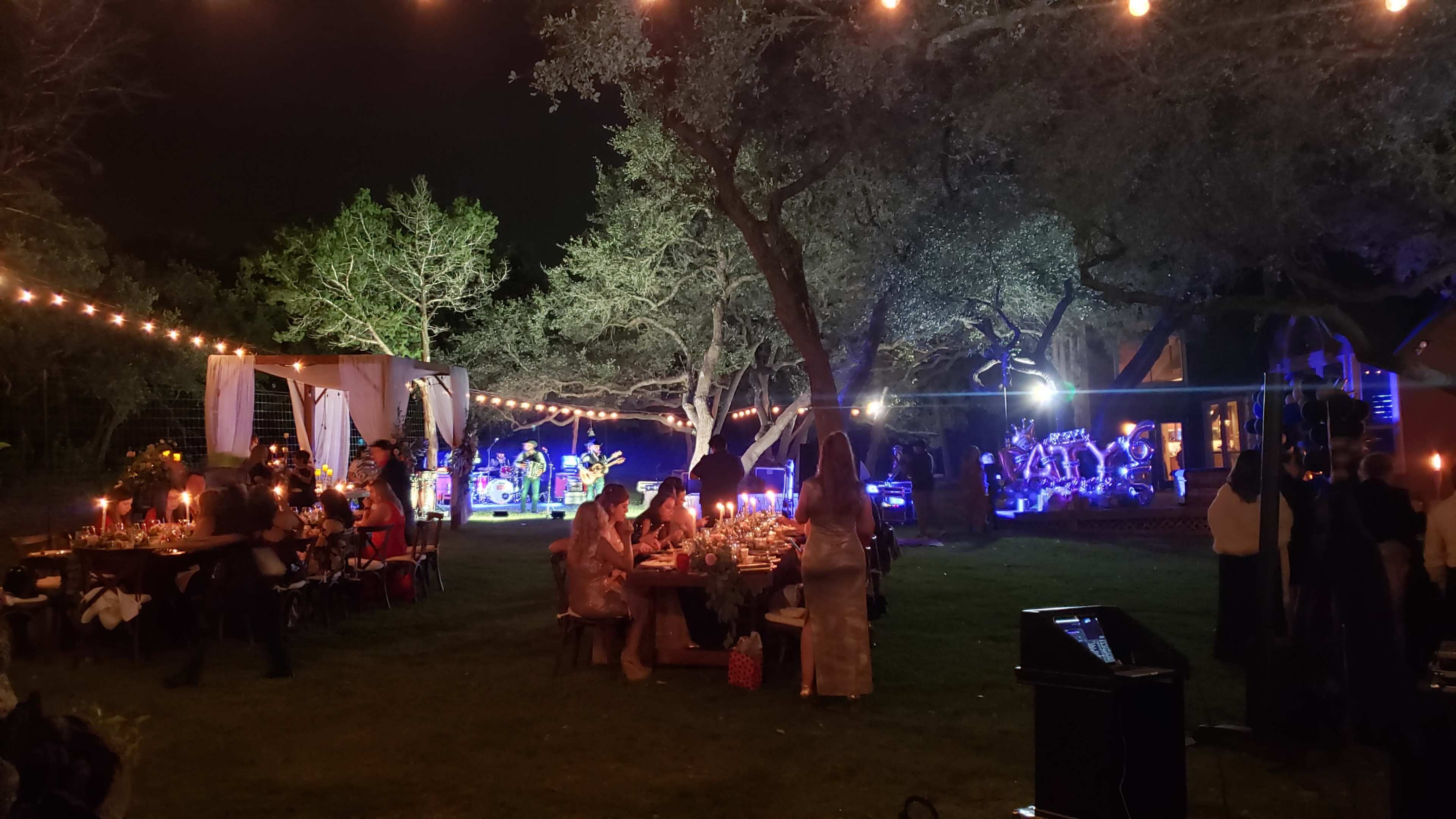 A lively outdoor evening gathering features a band playing music under string lights, surrounded by tables of guests enjoying the festive atmosphere.