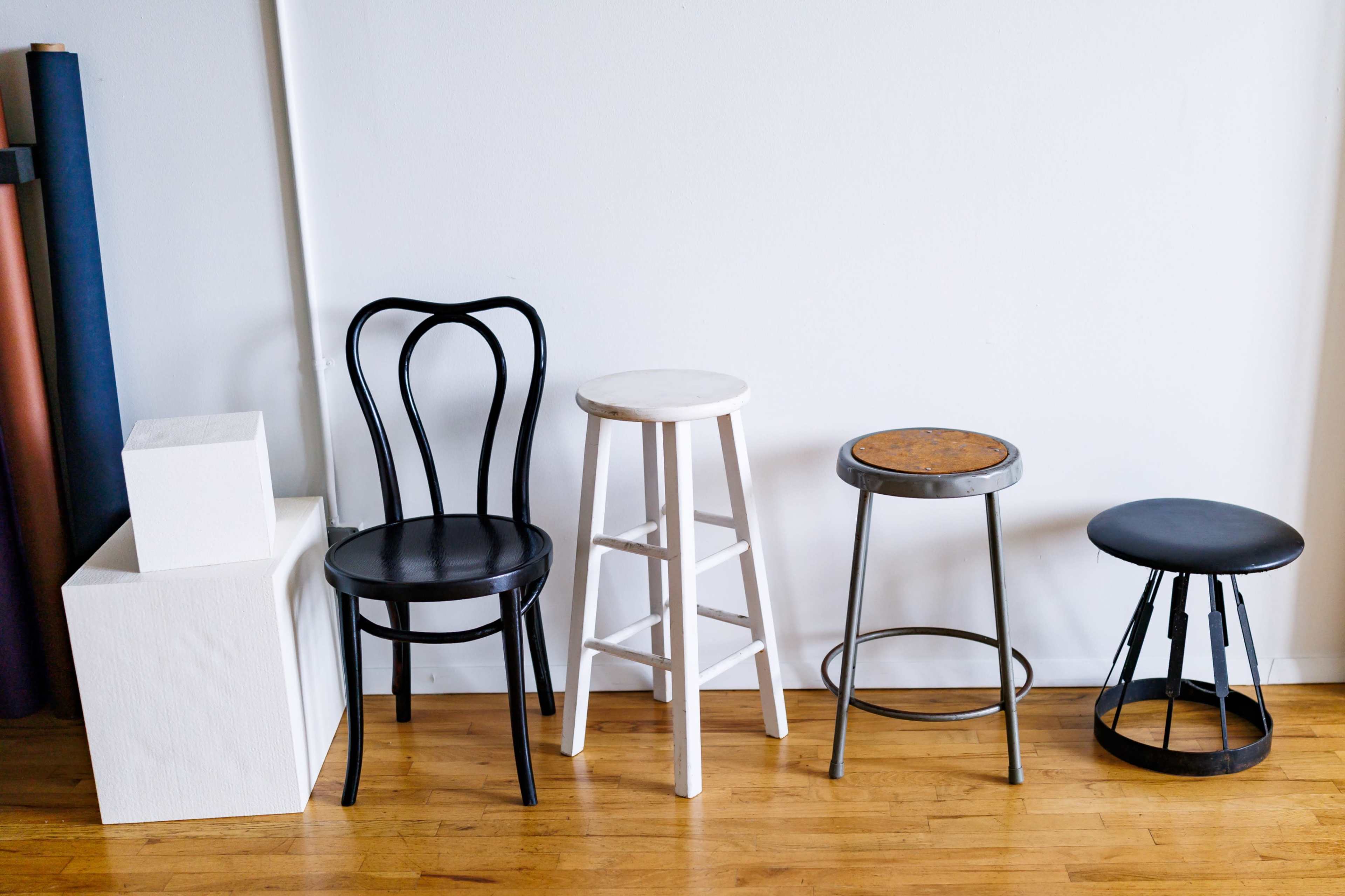 The image shows four different types of stools and a black chair arranged next to a white cube on a wooden floor against a plain white wall.