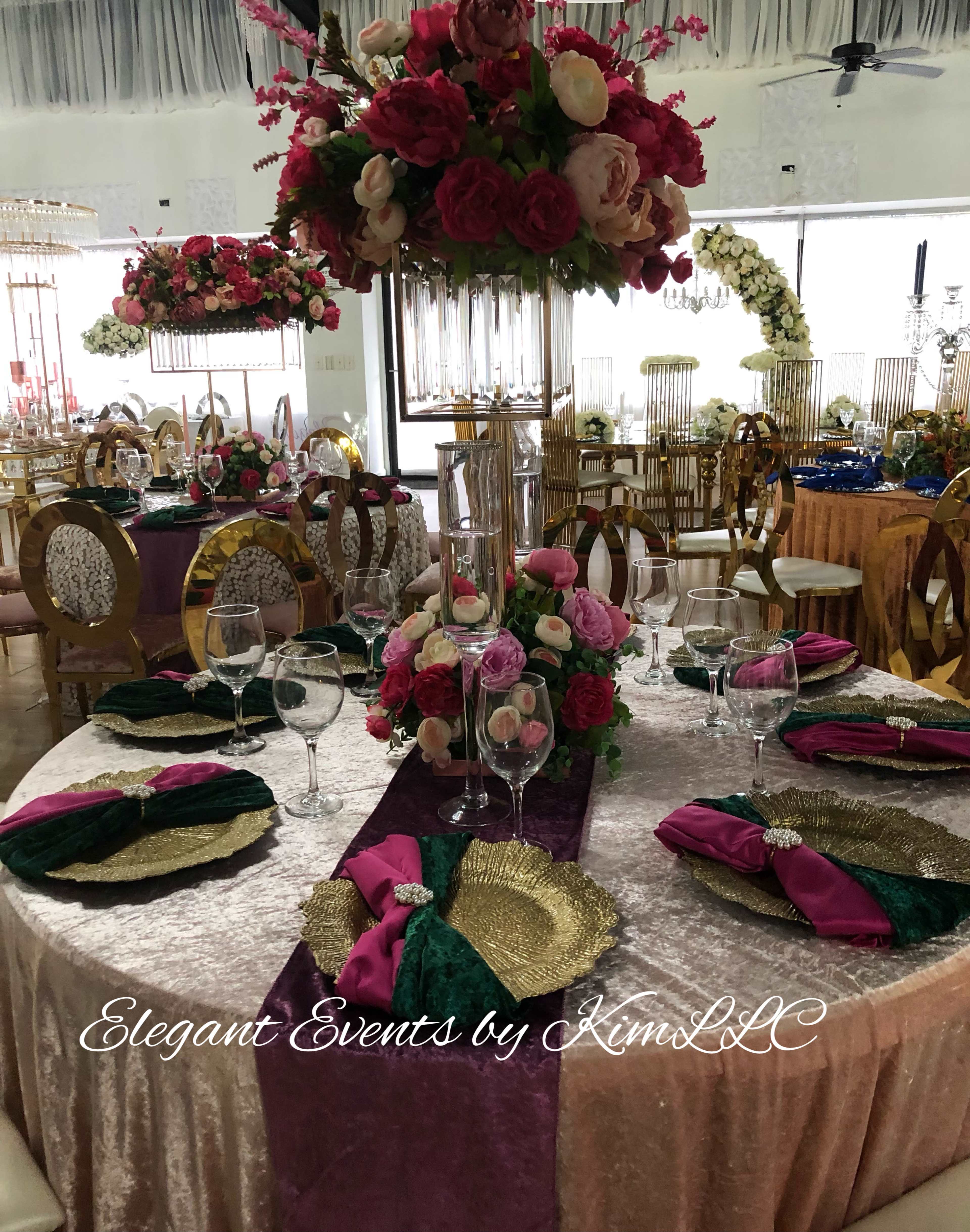 A decorated event space features a table set with elegant floral arrangements, fine dishware, and rich-colored linens.