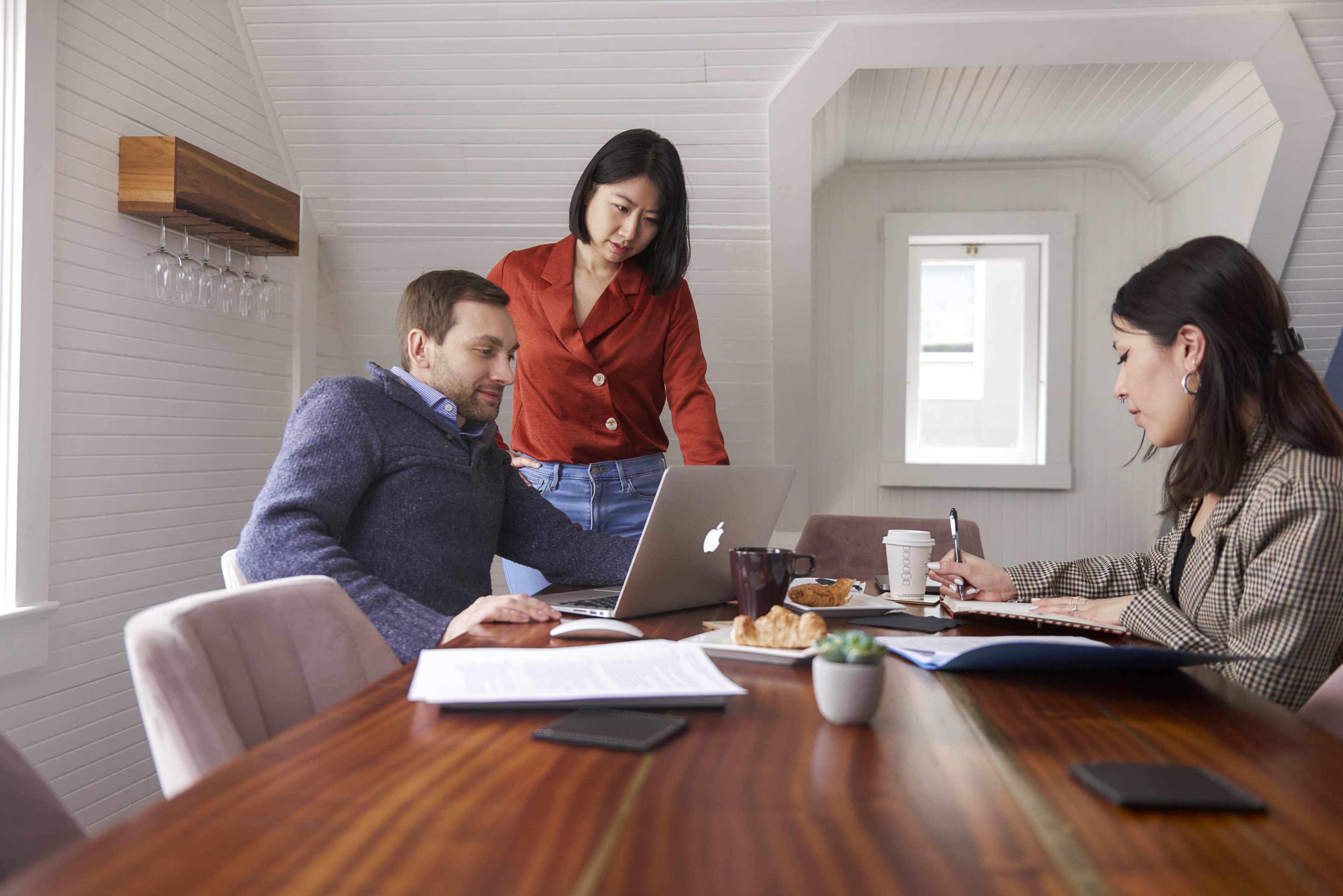 Three individuals are engaged in a collaborative work session at a table, with one person sitting at a laptop and the others discussing while surrounded by pastries and coffee.