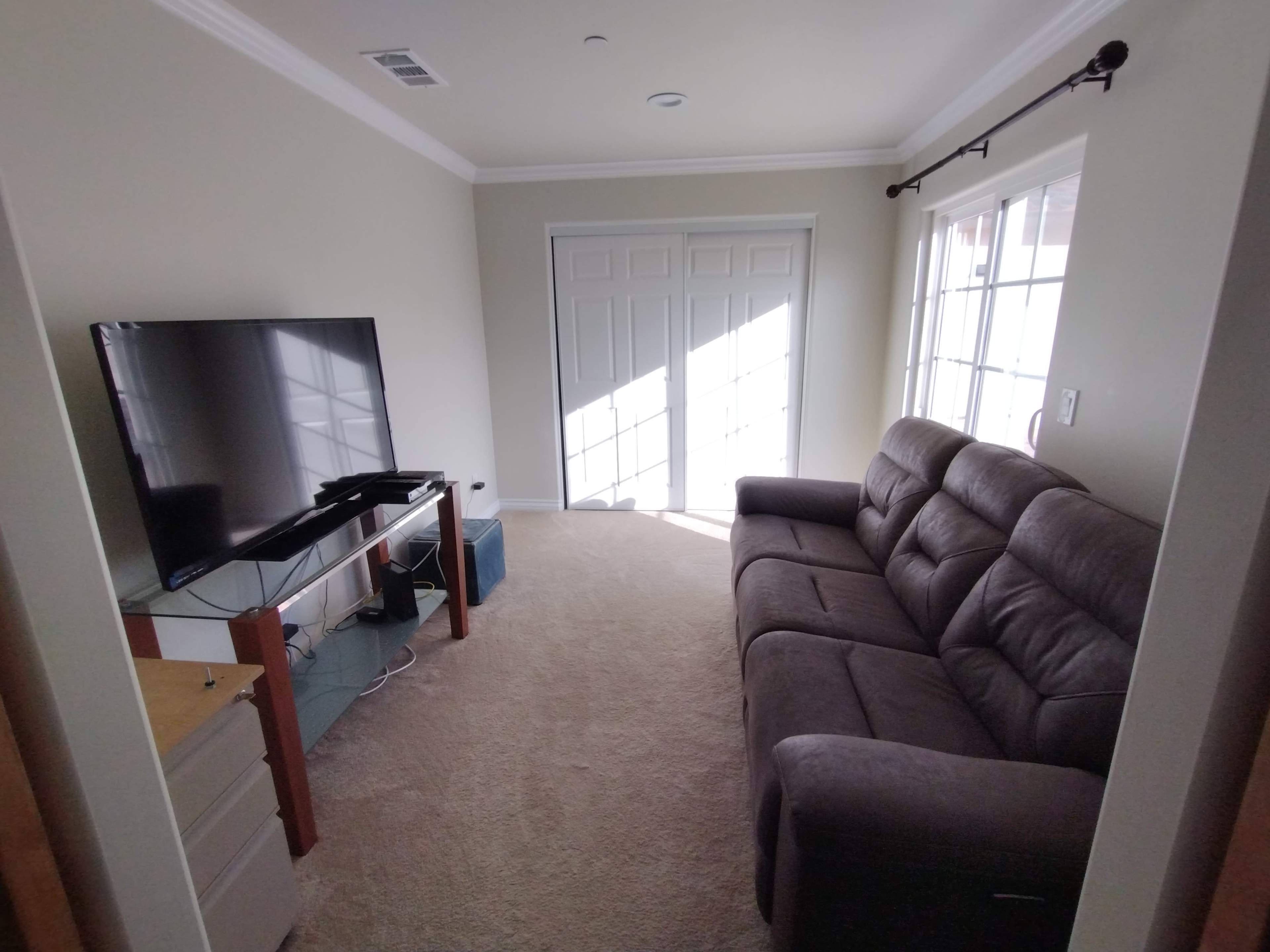 A living room features a gray sofa, a television on a stand, and a large window with light streaming in.