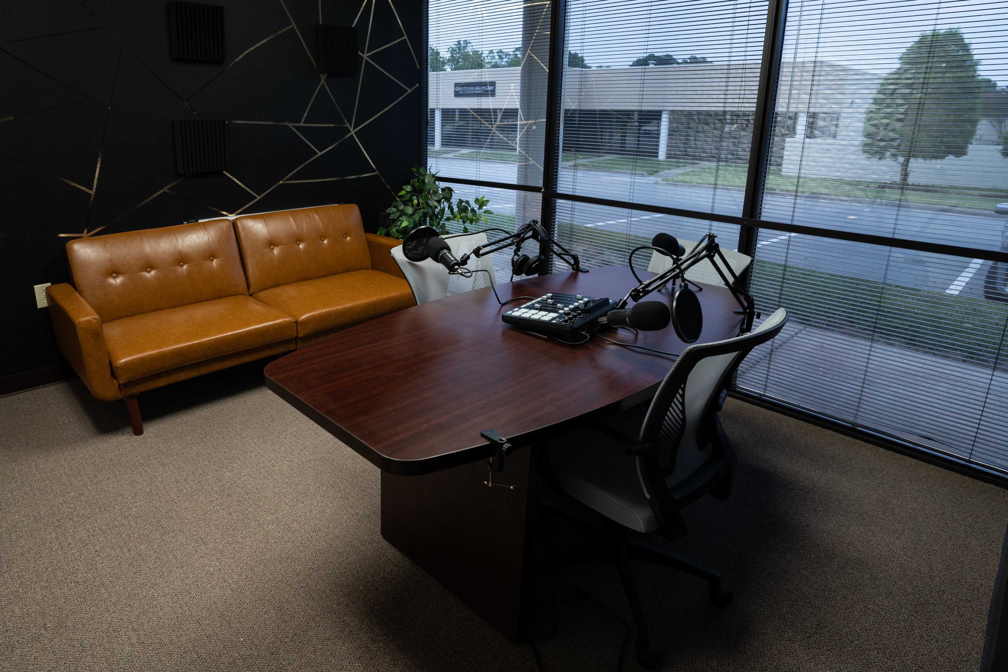 A small recording studio equipped with a brown sofa, a table with microphones, and two office chairs, located near a window with blinds.